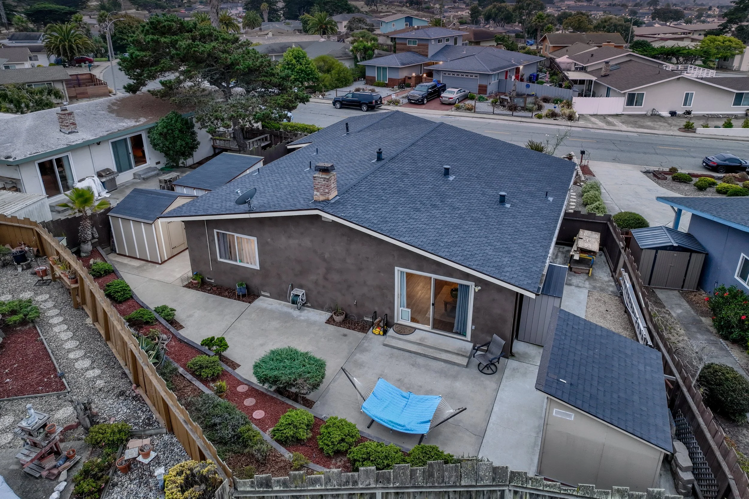 Aerial view of a backyard with a concrete patio, small garden beds, and a blue hammock. The house has gray walls, a dark shingle roof, and a sliding glass door. Surrounding the yard are neighboring houses, trees, and a street in Seaside, California.