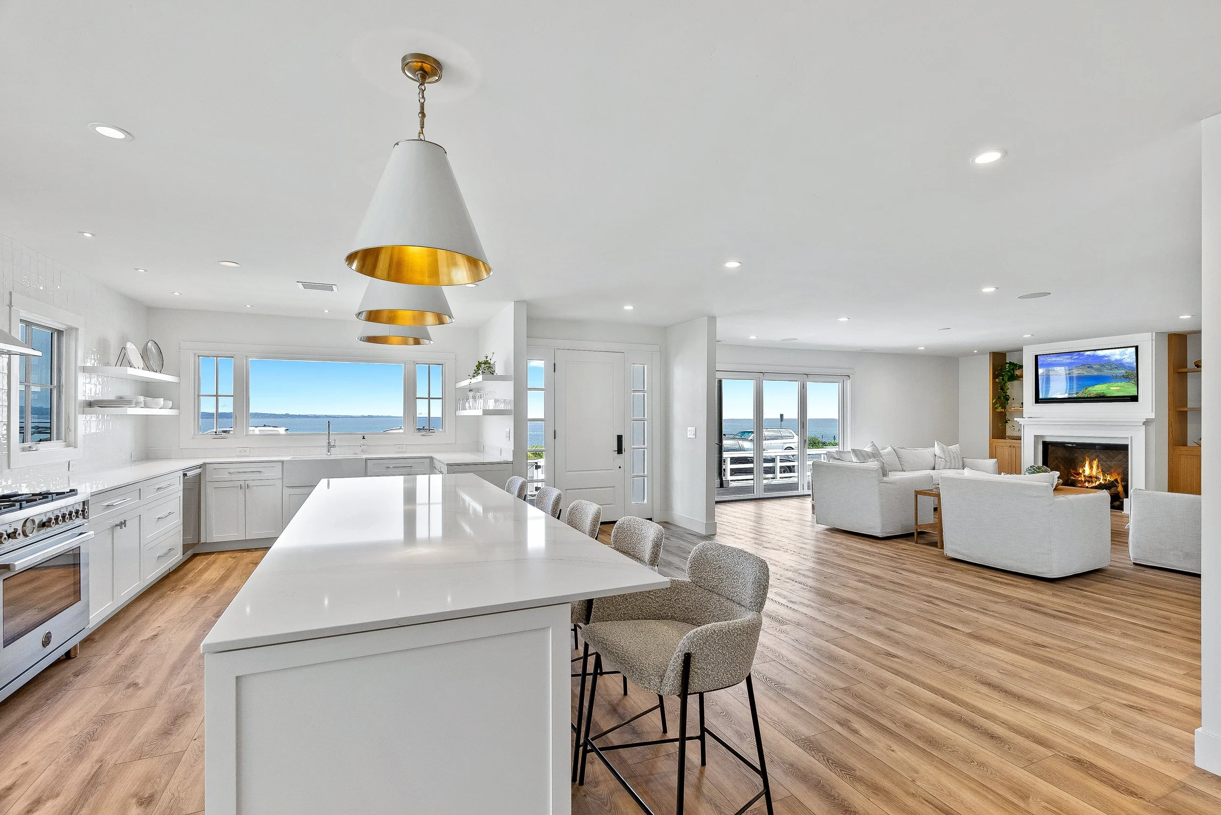 Open-concept living room and kitchen with large windows overlooking water, white furniture, hardwood floors, fireplace with TV, and sliding glass doors to balcony.