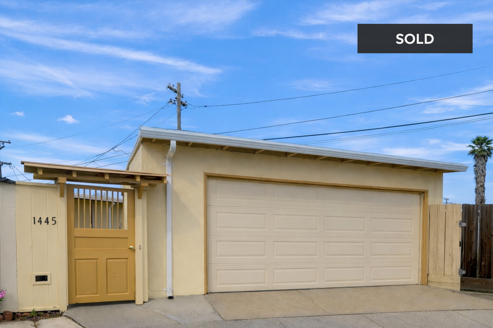 A yellow garage with a matching side gate, house number 1445, under a blue sky with wispy clouds, and a