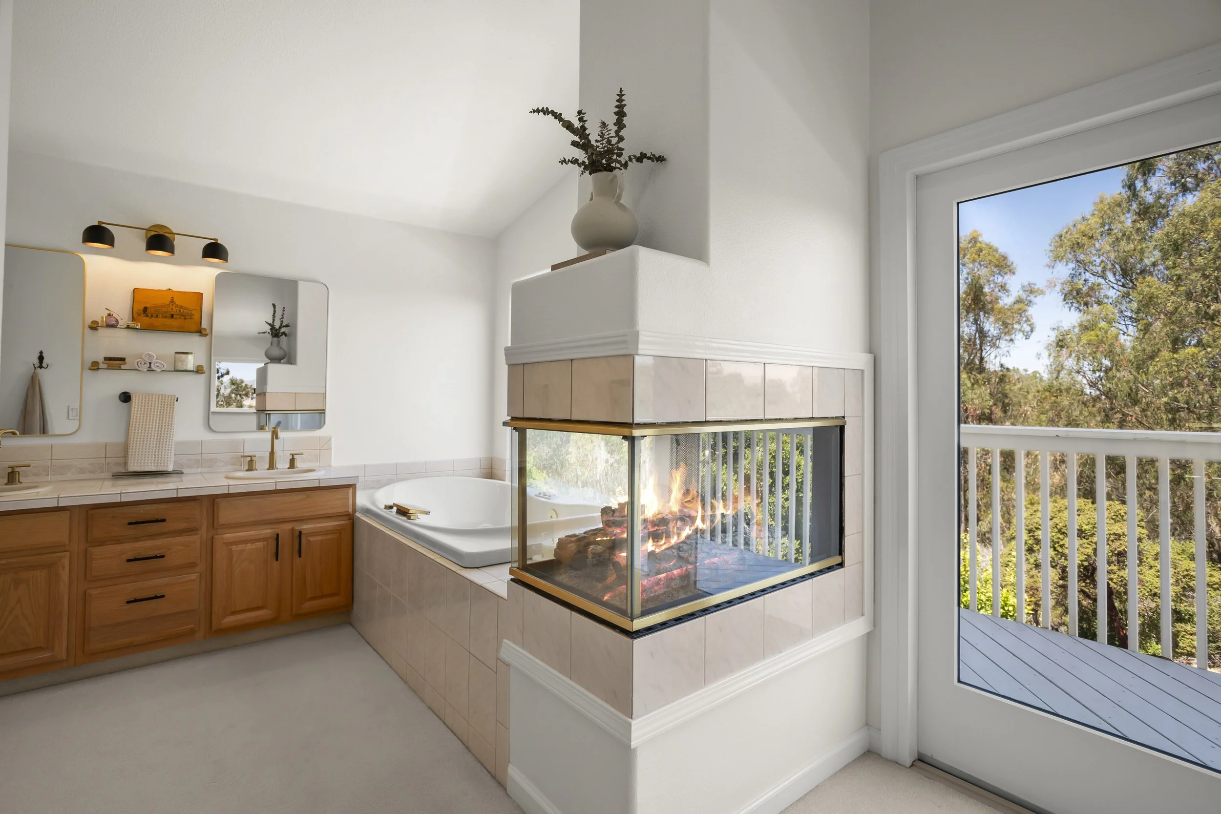 A bathroom with a corner fireplace, a bathtub, a vanity with double sinks, and a sliding glass door leading to a balcony with trees outside.