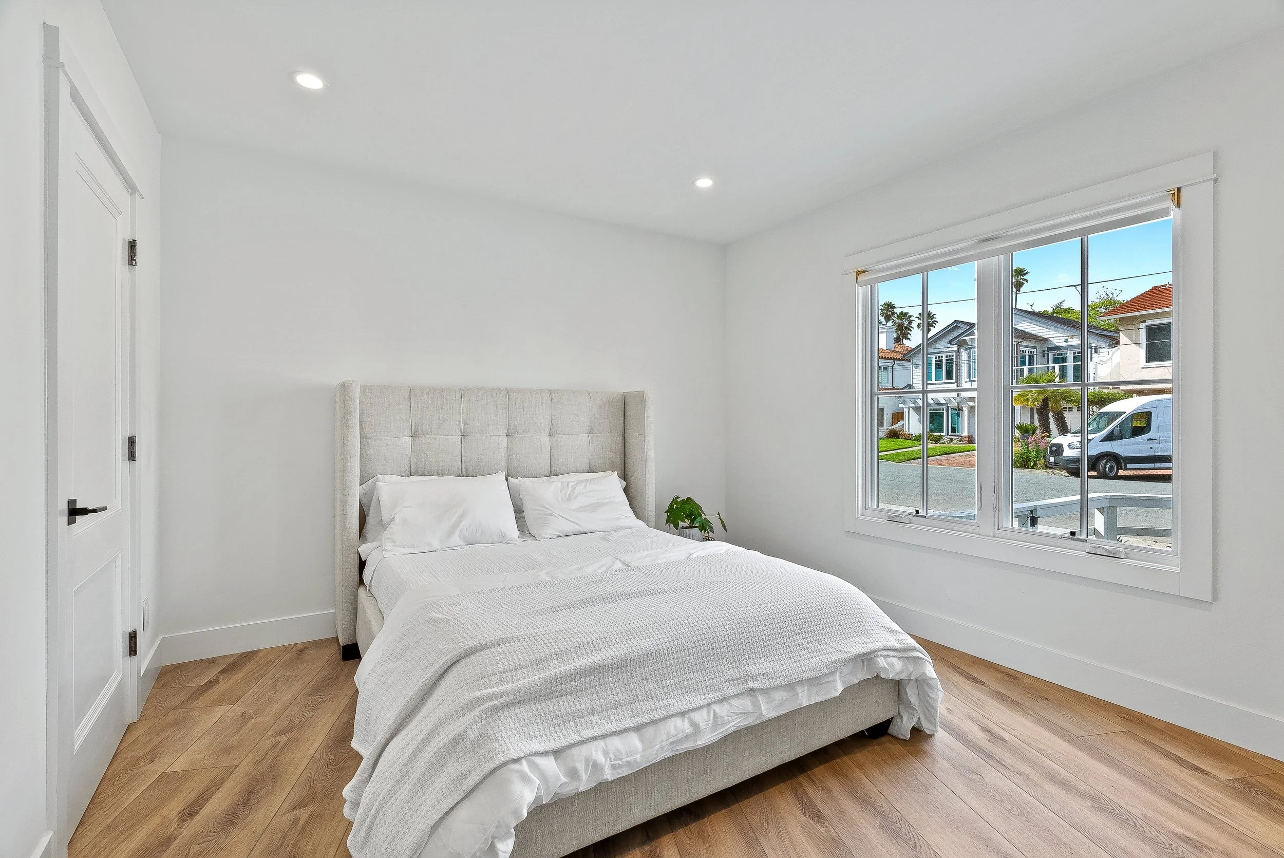 A clean, minimalist bedroom with a white bed, white walls, and a large window showing a street with houses and palm trees outside in Capitola, California.