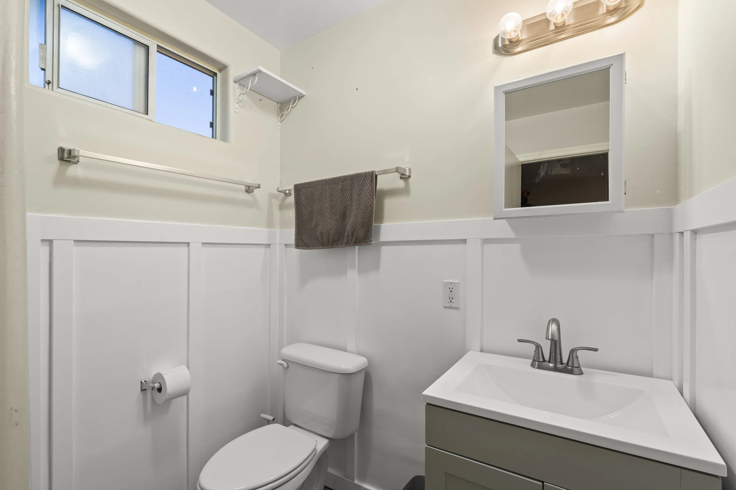 Small bathroom with a window, towel rack with a brown towel, wall-mounted shelf, mirror, sink with a gray vanity, toilet, and wall outlet.
