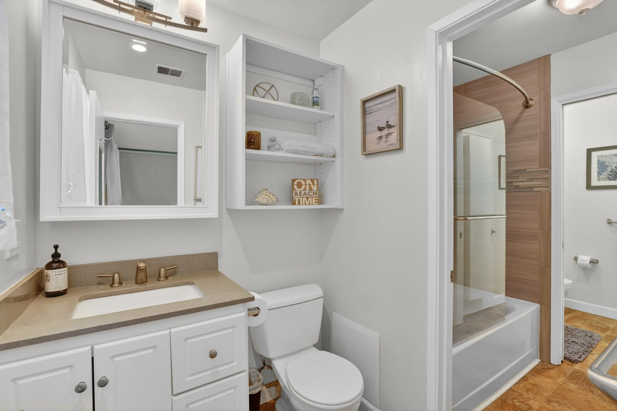 Bathroom with white vanity, beige countertop, mirror, toilet, open shelving, and shower with glass door and wooden accents.