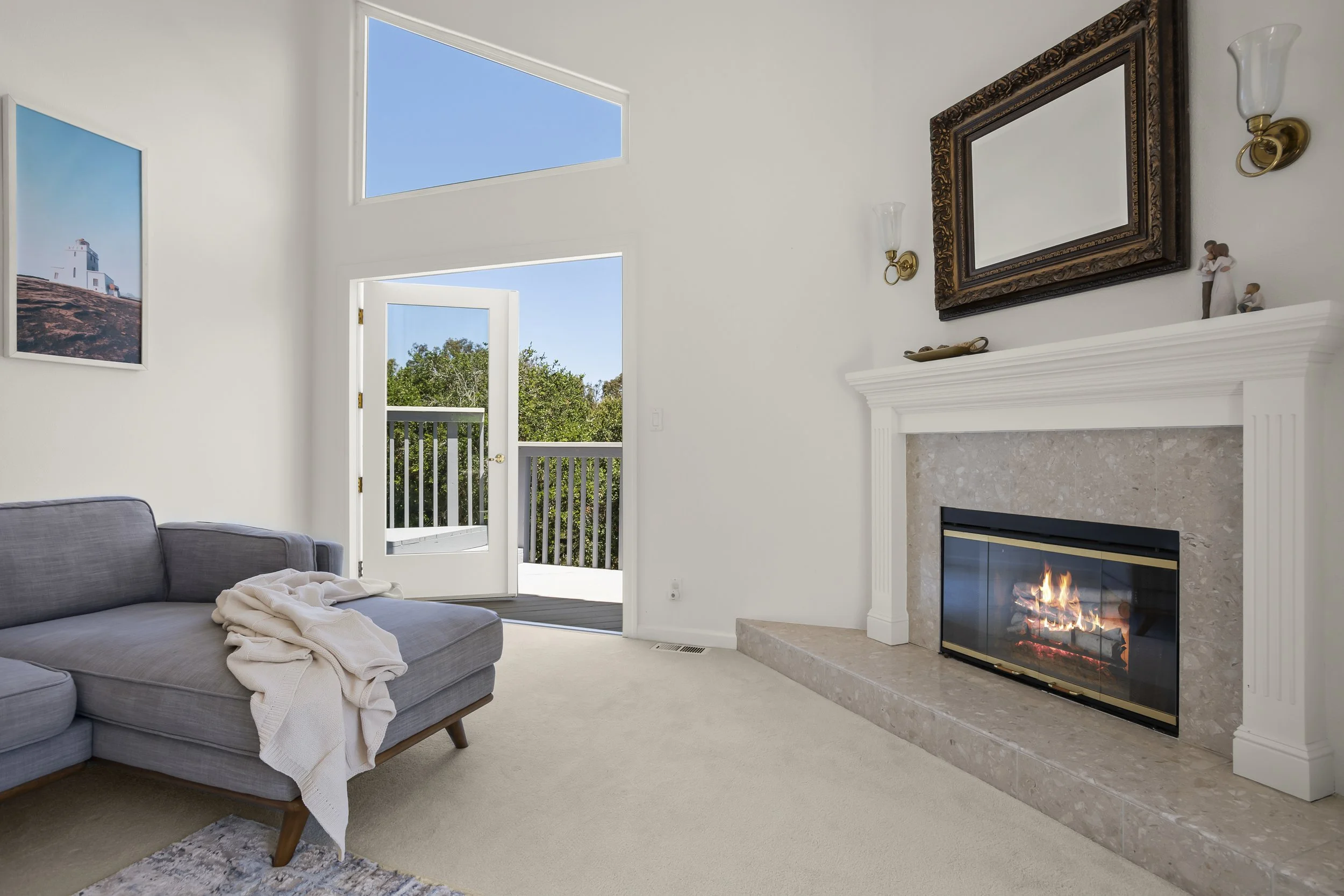 Living room with a gray couch, a fireplace with a fire, a mirror above the fireplace, a pair of wall sconces, a painting on the wall, and an open door leading to a balcony with trees outside.