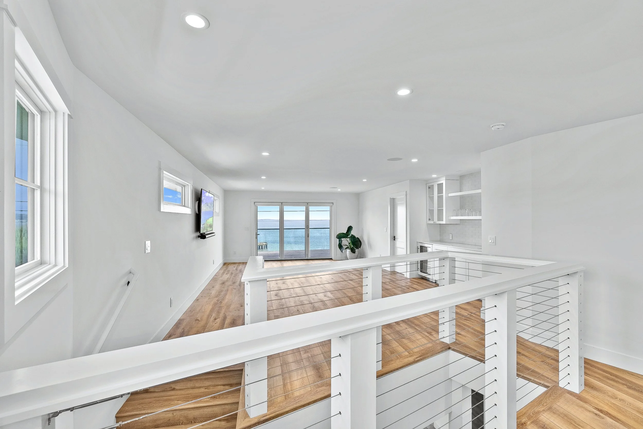 Living room with white walls, wooden floors, large windows, a flat-screen TV, and a glass door leading to an ocean view in Capitola, California.