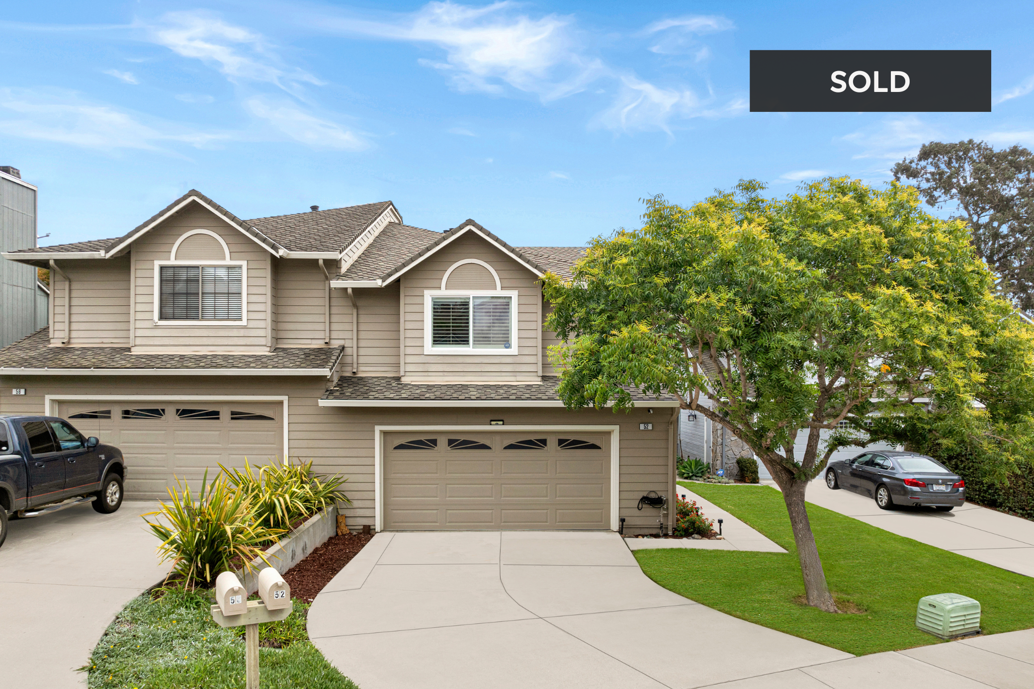 Front view of a two-story house with a beige exterior, two garages, a tree, and a lawn. A black truck is parked on the driveway, and a woman is walking on the sidewalk. A 'sold' sign is in the top right corner of the image 52 Winding Way Watsonville