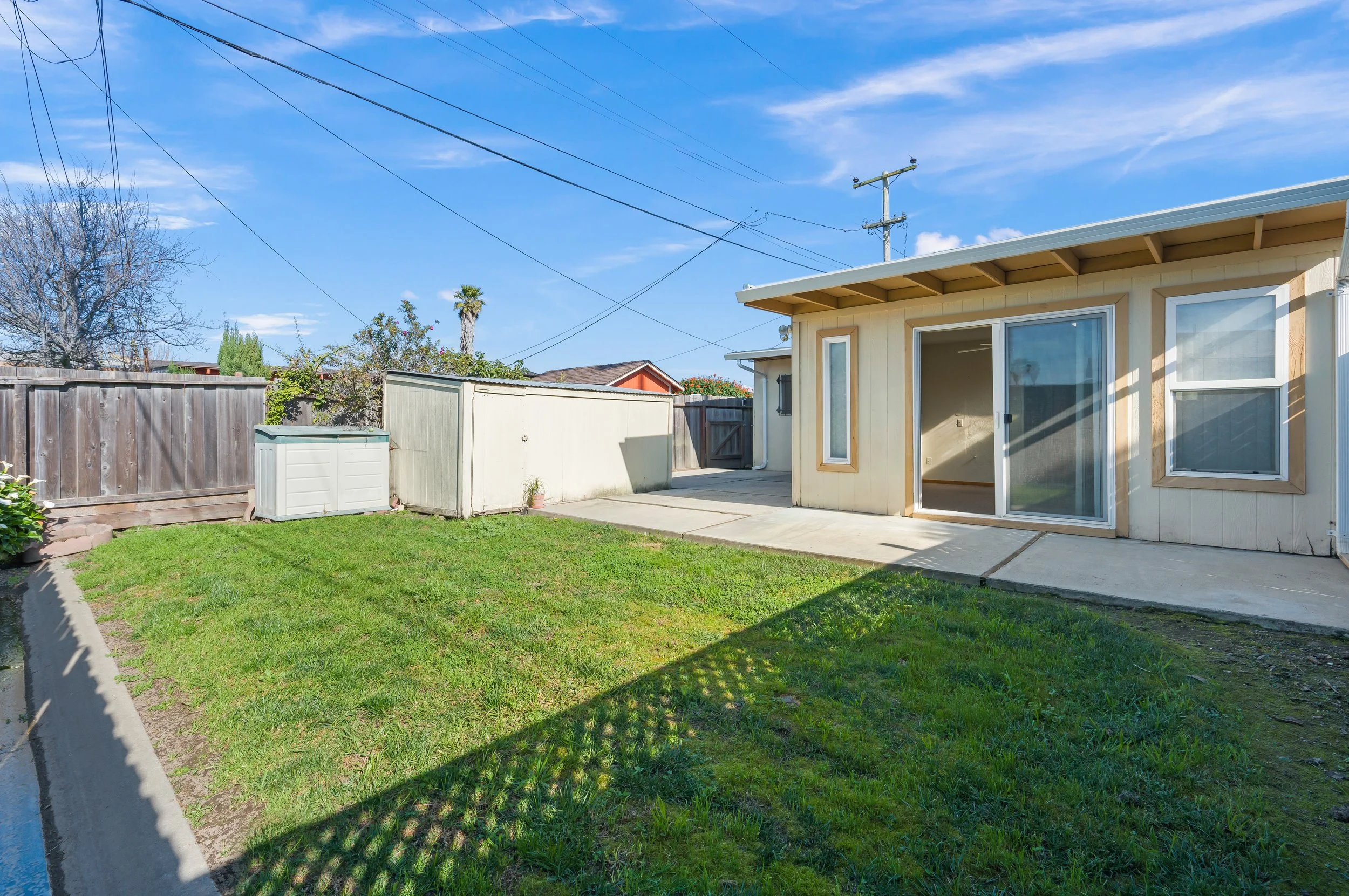 Backyard with green grass, concrete patio, shed, wooden fence, house with sliding glass door, and sky with clouds in Salinas, Monterey county, CA.