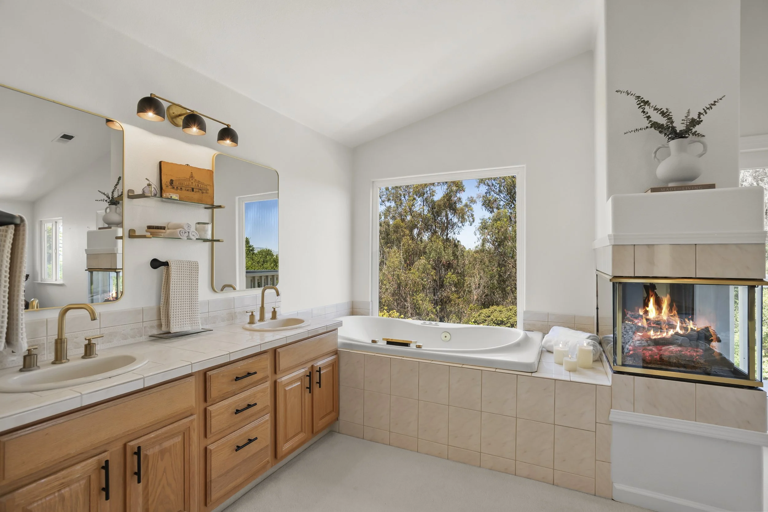 Bathroom with twin sinks, large window, double-sided fireplace, and a large bathtub near the window with trees outside.