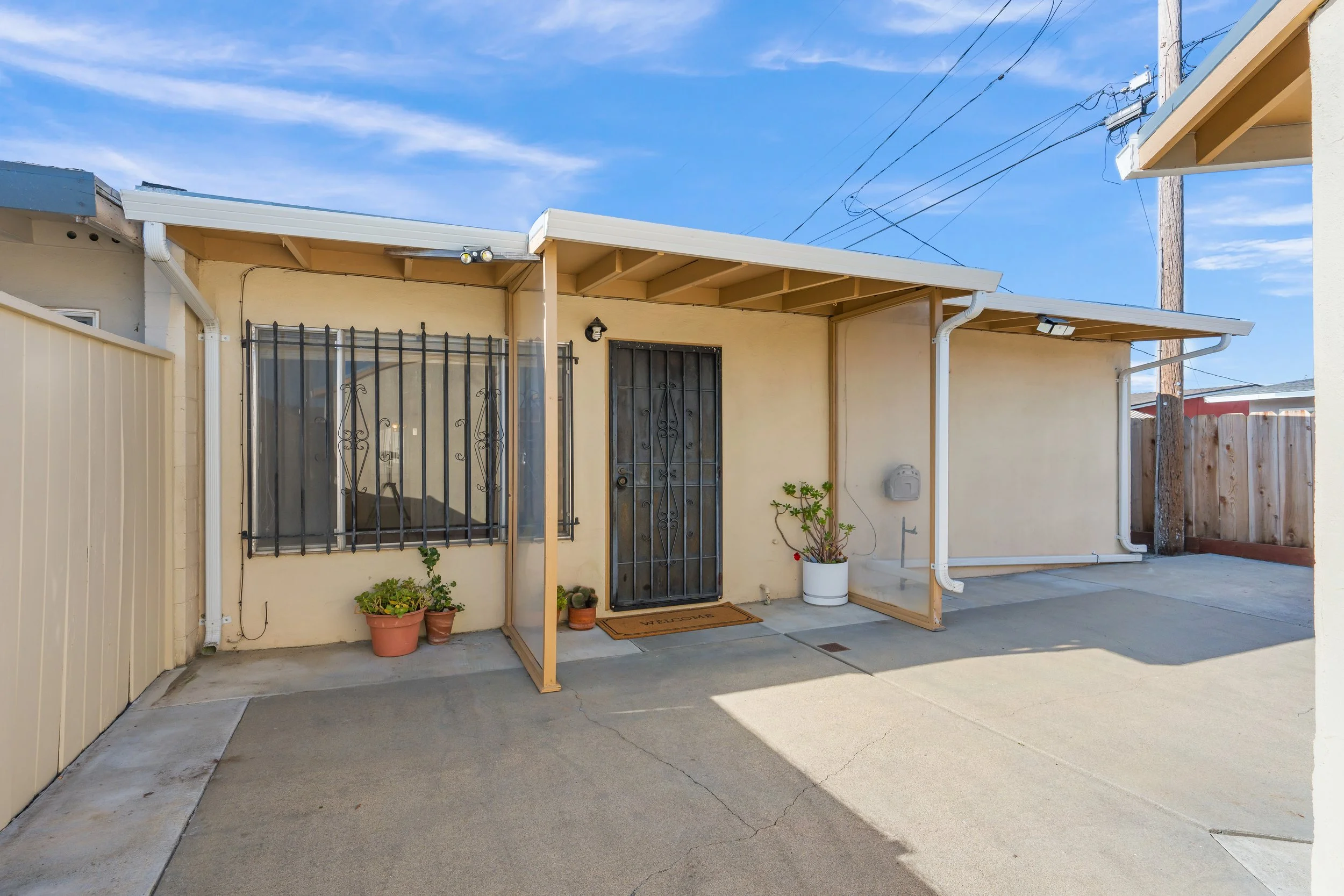 Backyard patio with a covered area, potted plants, a metal security door, and a window with bars, under a blue sky in Salinas, Monterey county, CA.