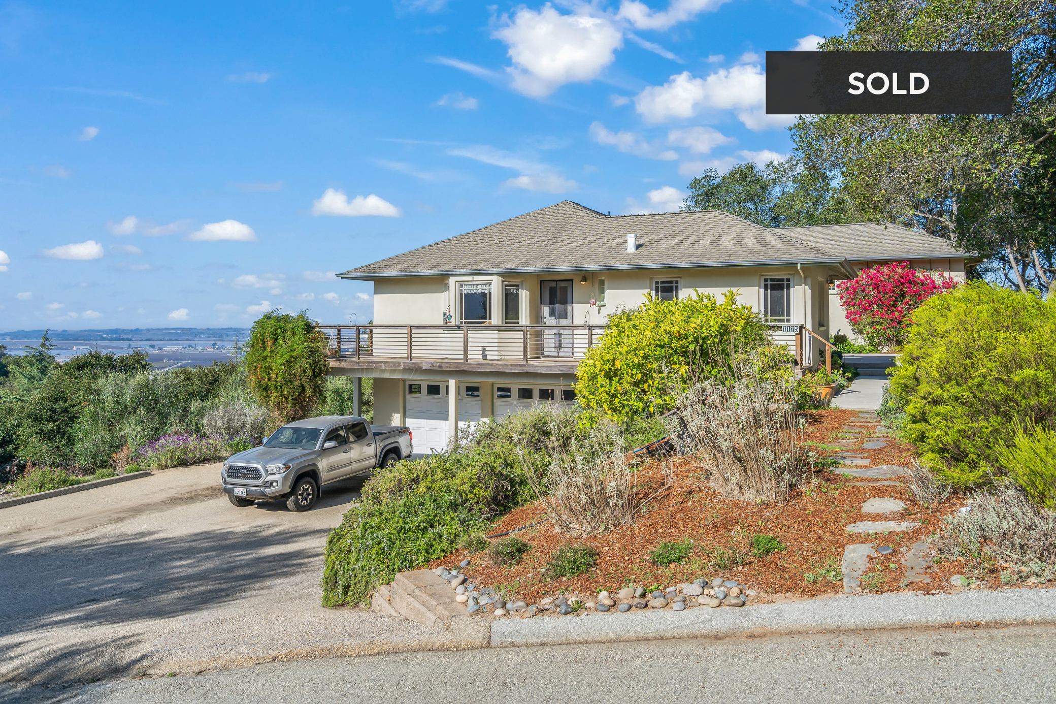 A house on a hill with a garage, a work truck parked in front on a gravel driveway, lush landscaping, colorful flowering bushes, and a blue sky with scattered clouds. The house has a balcony and is marked as sold.