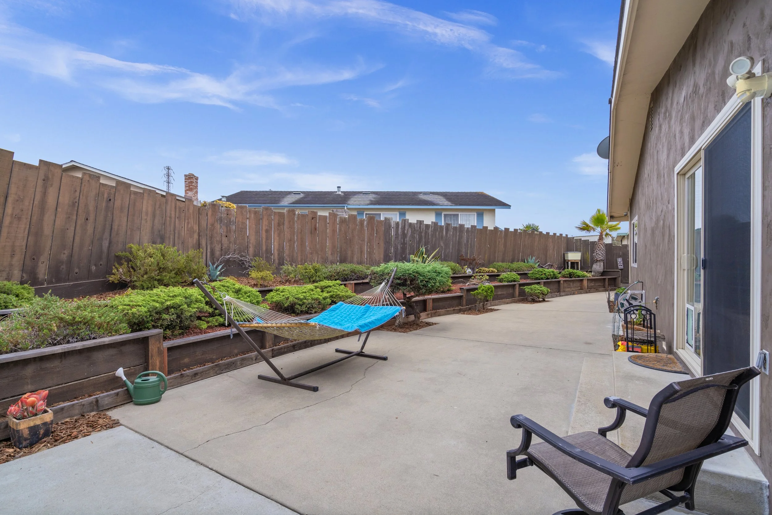 Backyard patio with a hammock, garden beds with plants, a watering can, a garden chair, and a sliding glass door, enclosed by a wooden fence, under a partly cloudy blue sky.