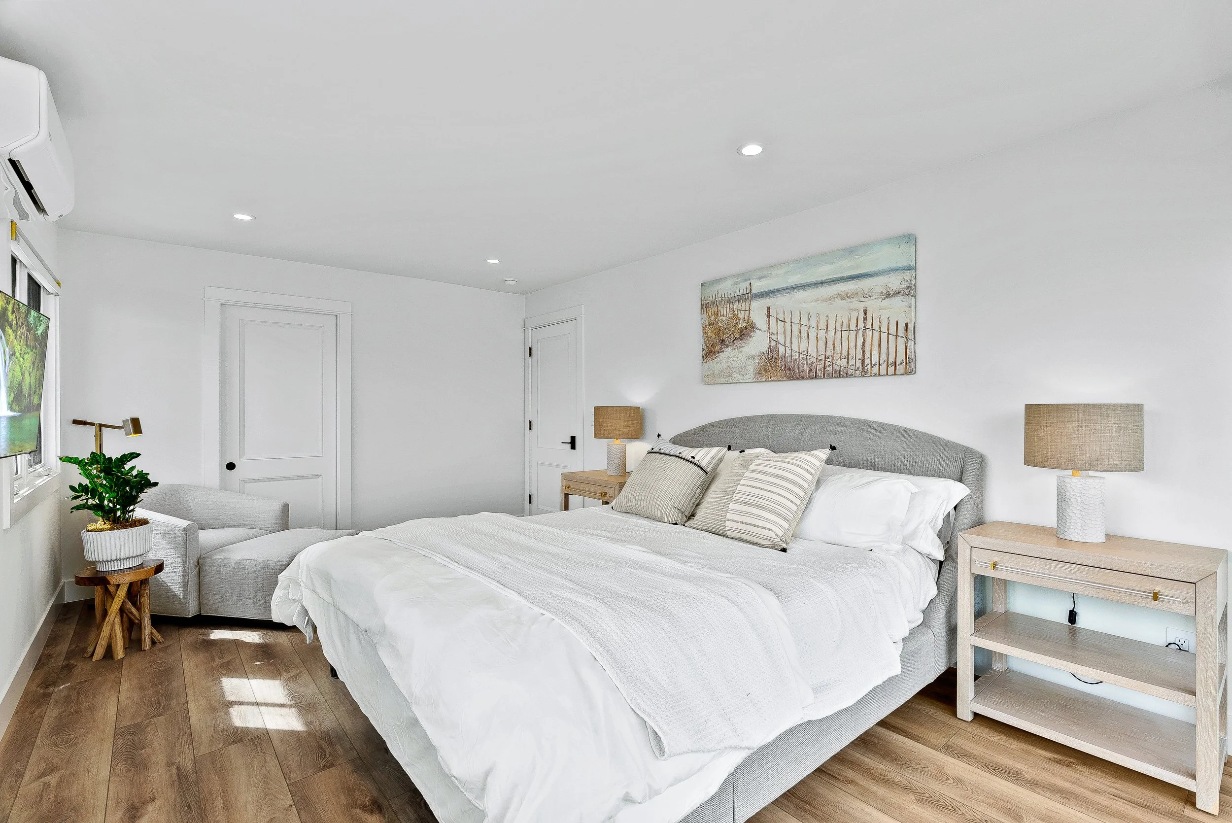 A bright and minimalist bedroom with a bed, nightstands, lamps, a sitting area with a plant, and a painting of a beach scene above the bed Capitola, California.