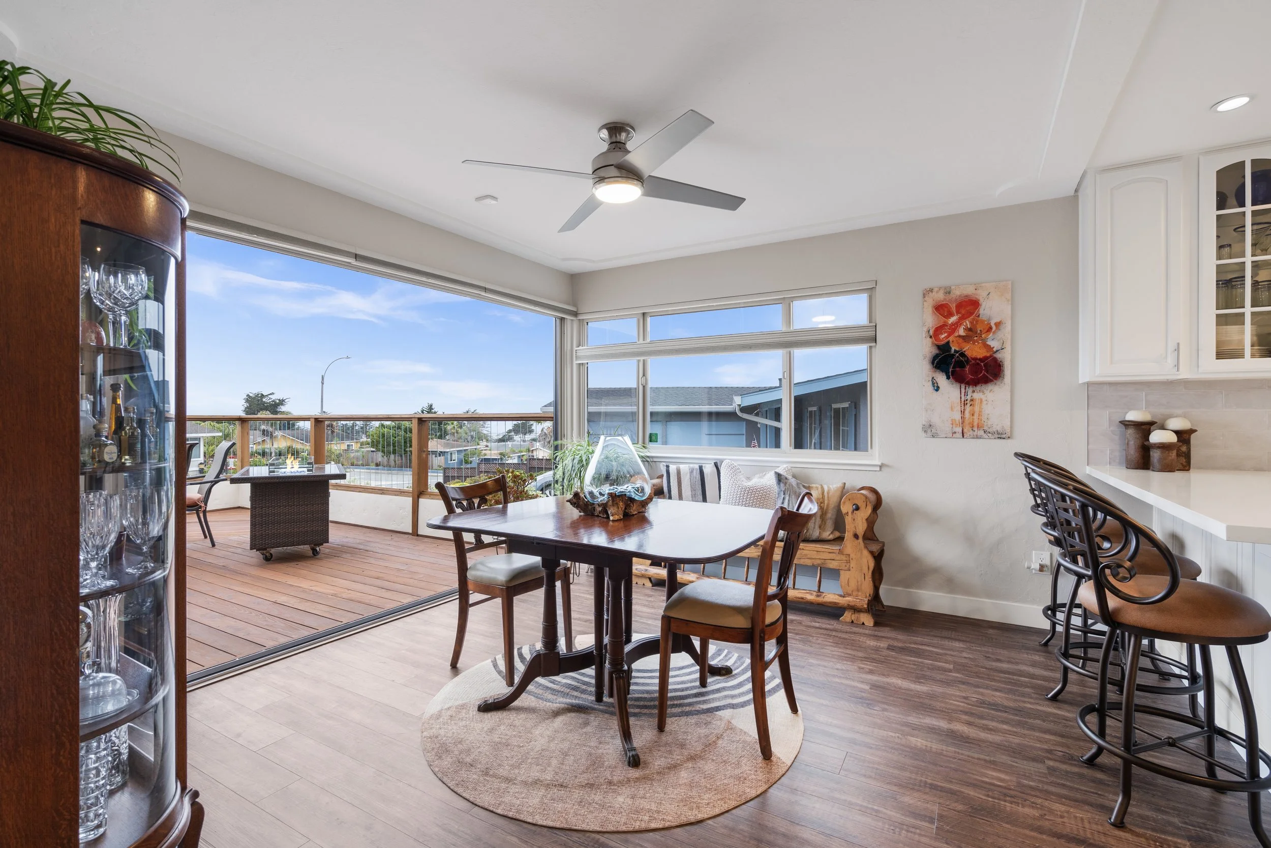 Open-concept dining and living area with a large glass sliding door leading to a wooden deck outside, wood flooring, a round rug under a wooden dining table with four chairs, a seating bench against the wall, a ceiling fan, and a bar with three stools.