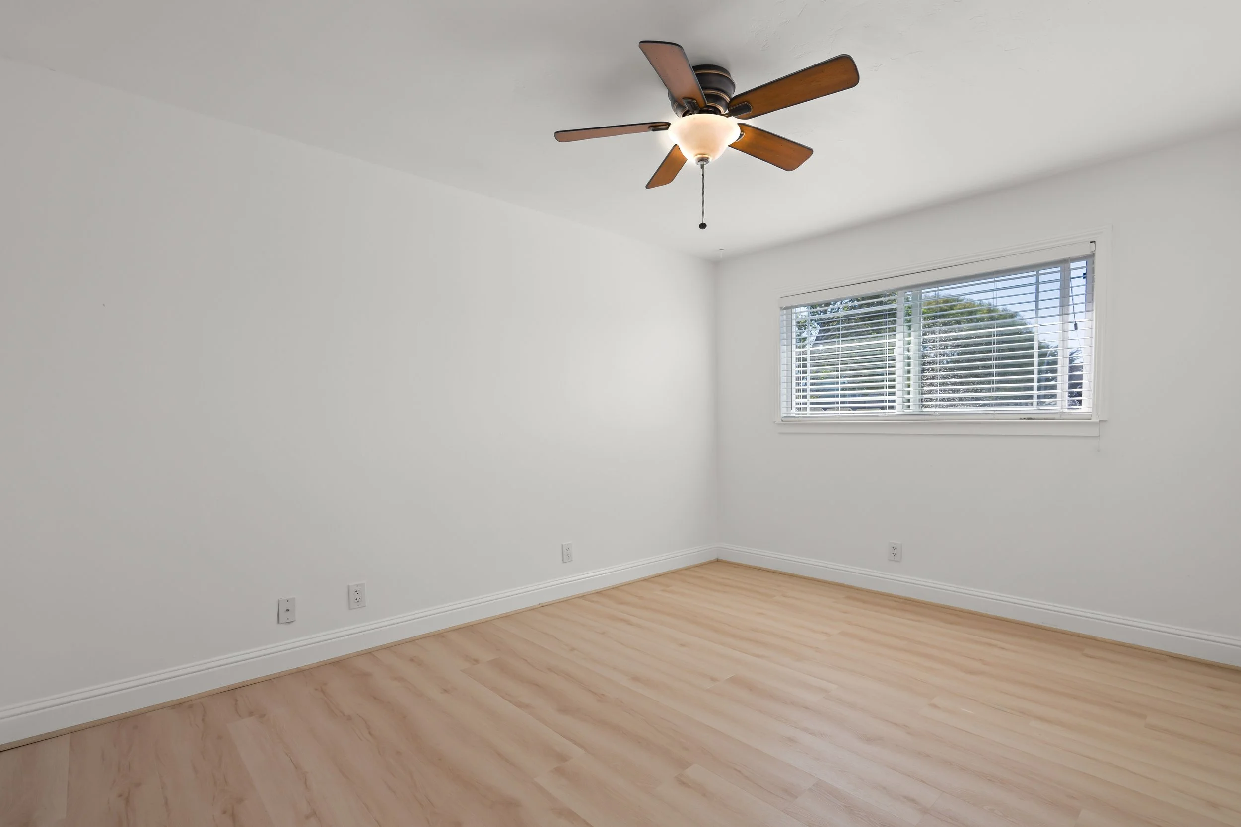 Empty room with white walls, hardwood floor, window with blinds, ceiling fan with light.
