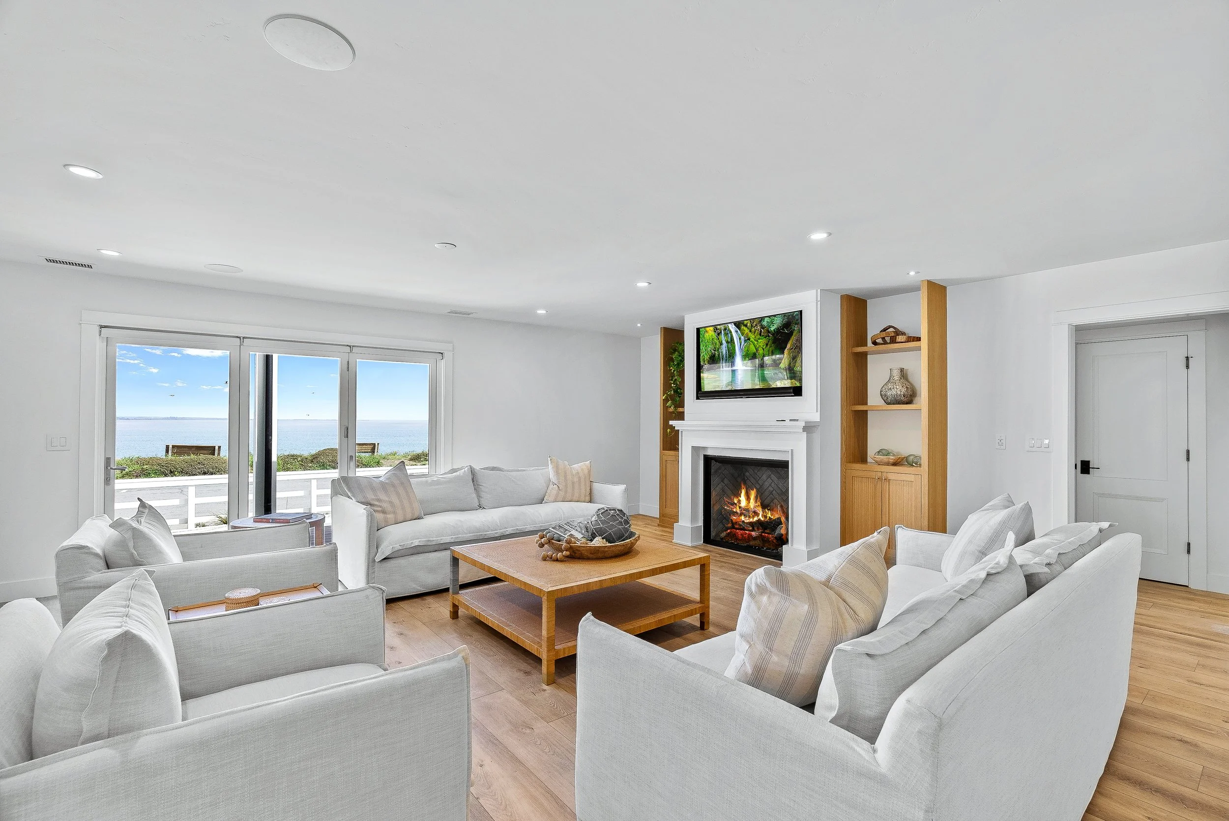 Bright living room with white sofas, wooden coffee table, fireplace, built-in shelves, and large sliding glass doors view of ocean in Capitola, California.