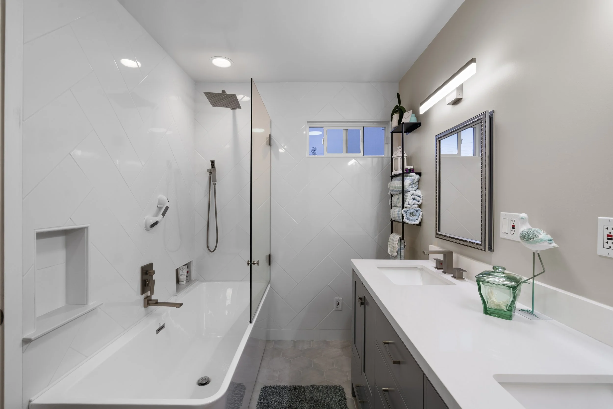 Modern bathroom with a bathtub and a shower on the left, a window above the bathtub, a vanity with a sink and mirror on the right, and shelves with folded towels and decorative items.