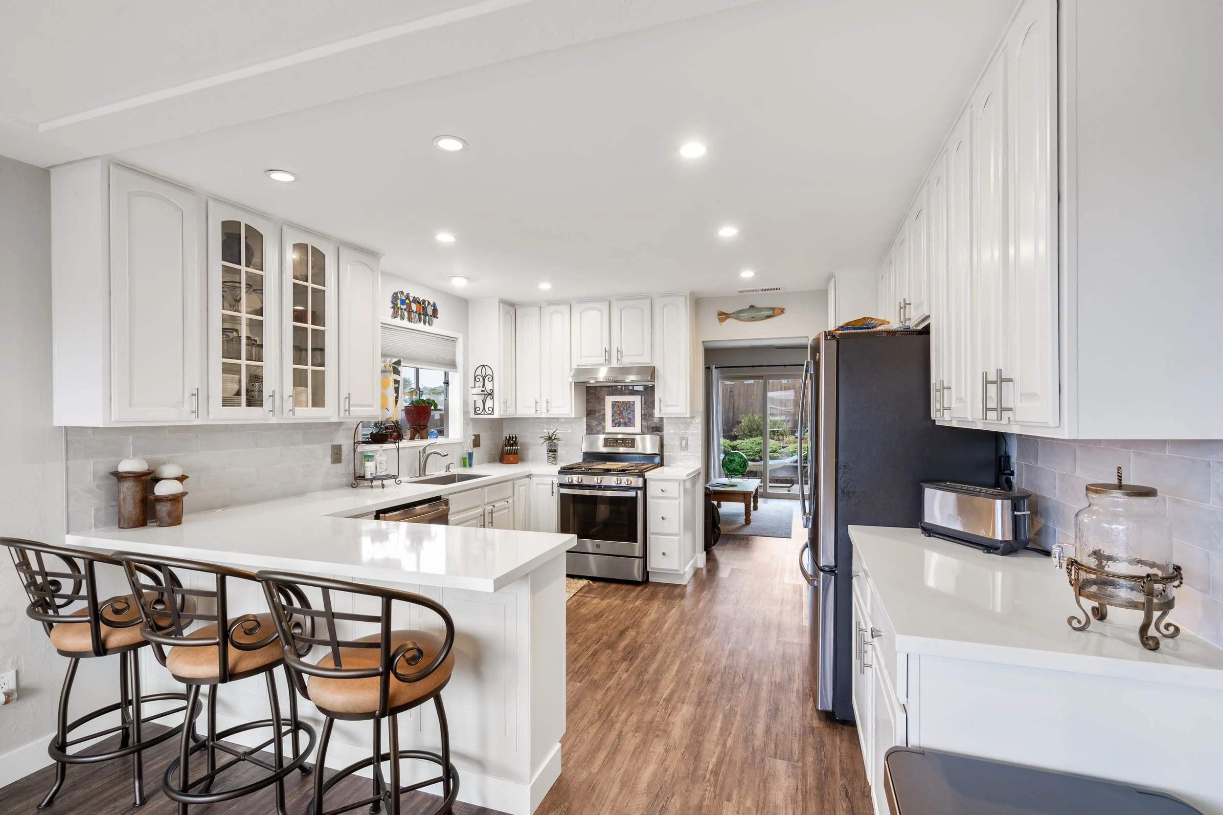 Bright kitchen with white cabinets, stainless steel appliances, a breakfast bar with three brown chairs, and hardwood flooring.