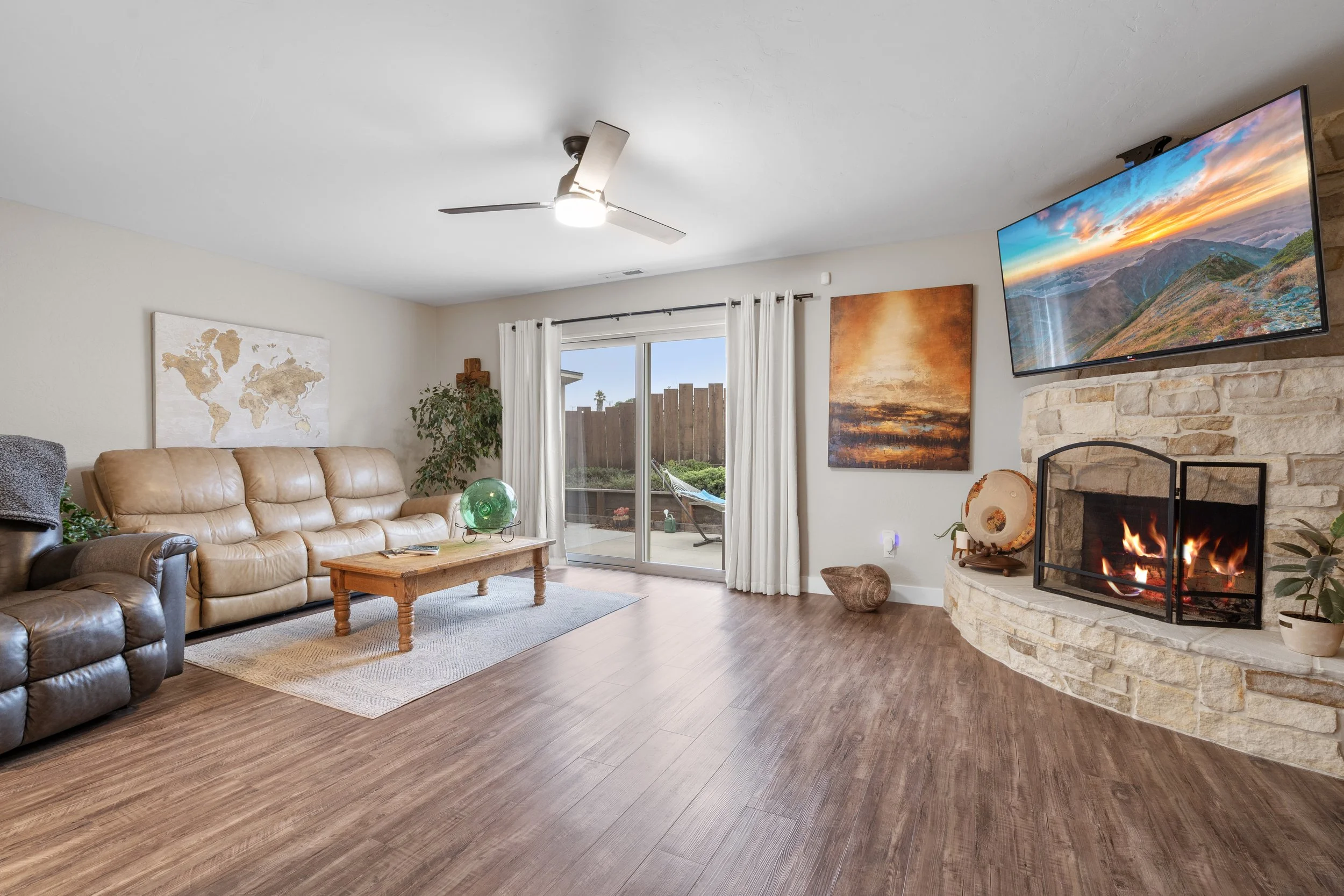 Living room with leather sofa, recliner, wooden coffee table, framed world map, sliding glass door with white curtains, fireplace with fire, wall-mounted TV, landscape paintings, plants, and wooden flooring.