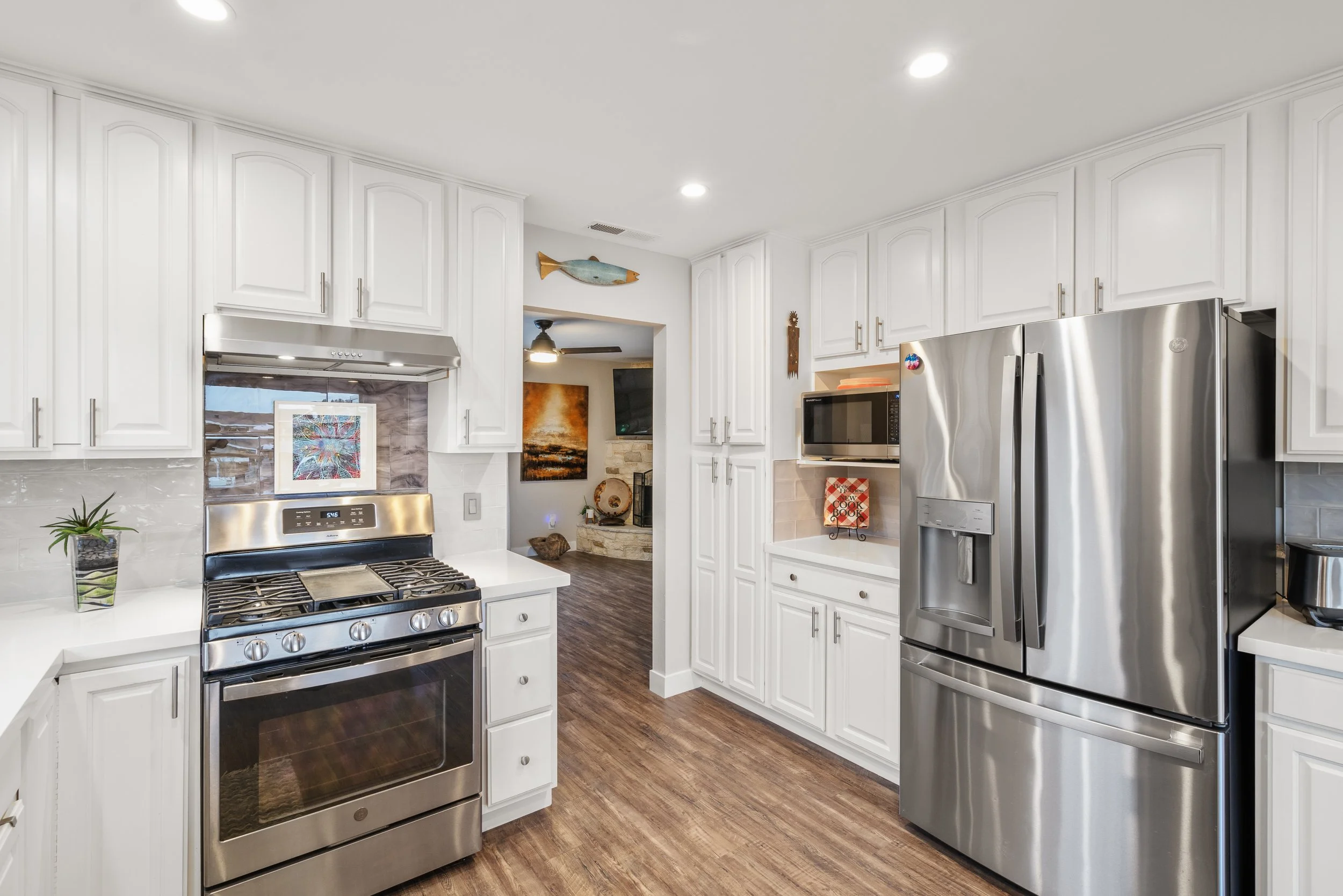 Modern white kitchen with stainless steel appliances, including a refrigerator, stove, and microwave. White cabinets, wood flooring, and a small plant on the counter.