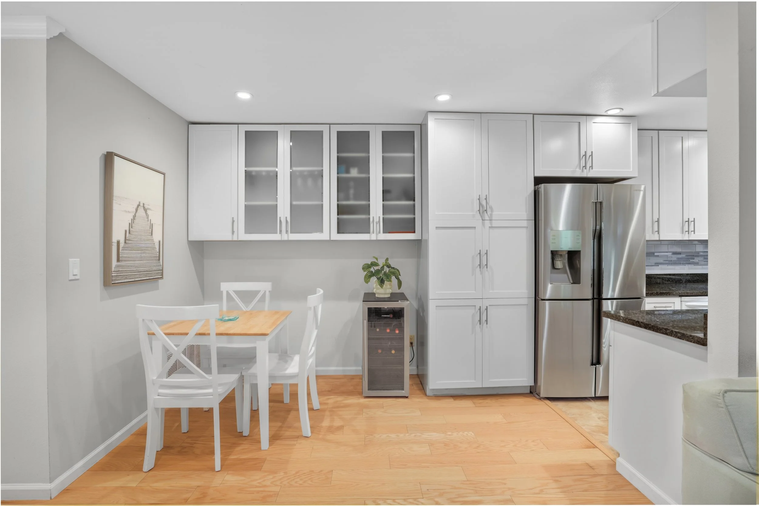 Kitchen with white cabinets, a stainless steel refrigerator, a small bar area, a dining table with four white chairs, a wine cooler, and a wall art of a pier extending into the water.