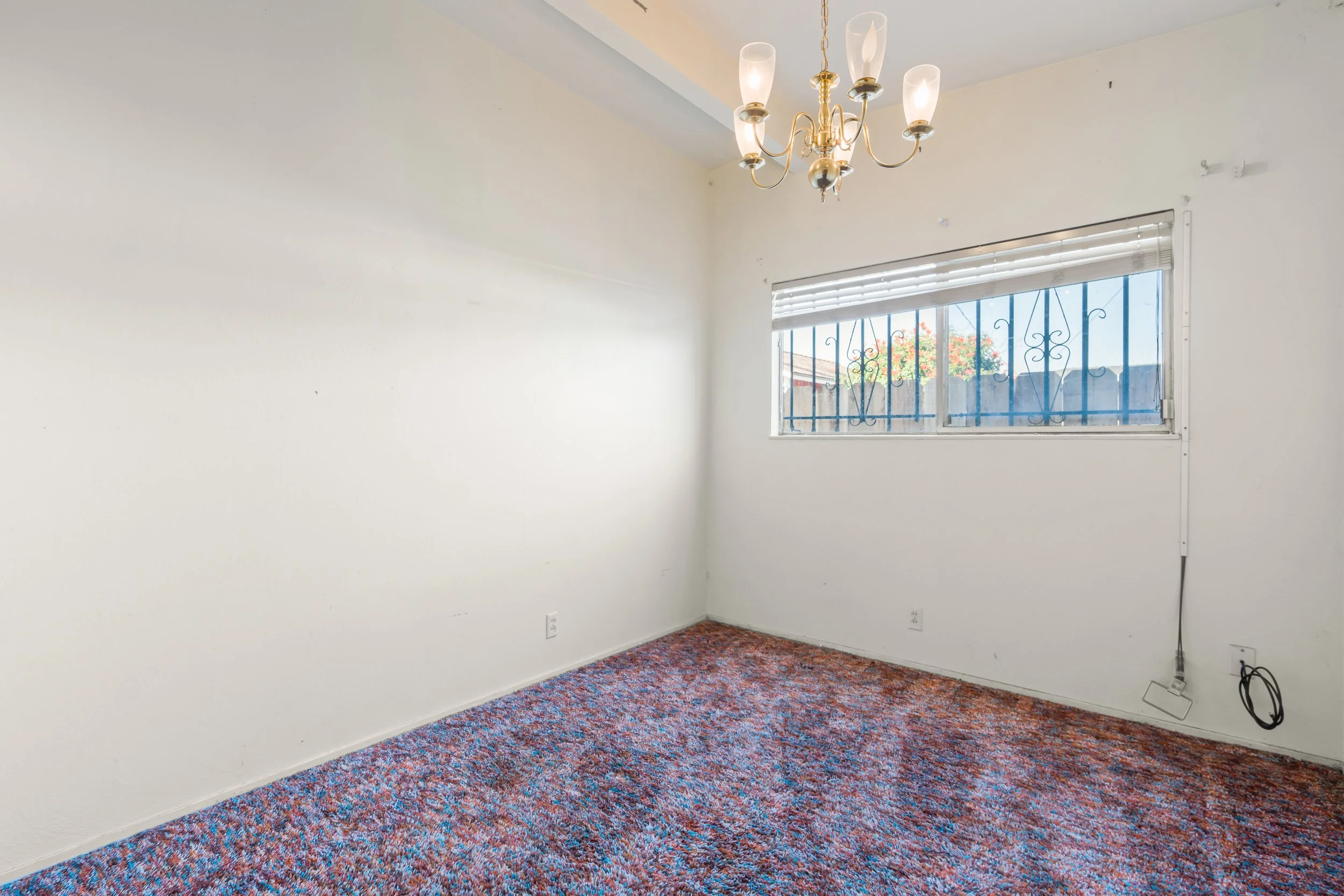 Empty room with white walls and colorful carpet, a window with metal bars, a chandelier hanging from the ceiling, and some electrical wiring near the outlet in Salinas, Monterey county, CA.