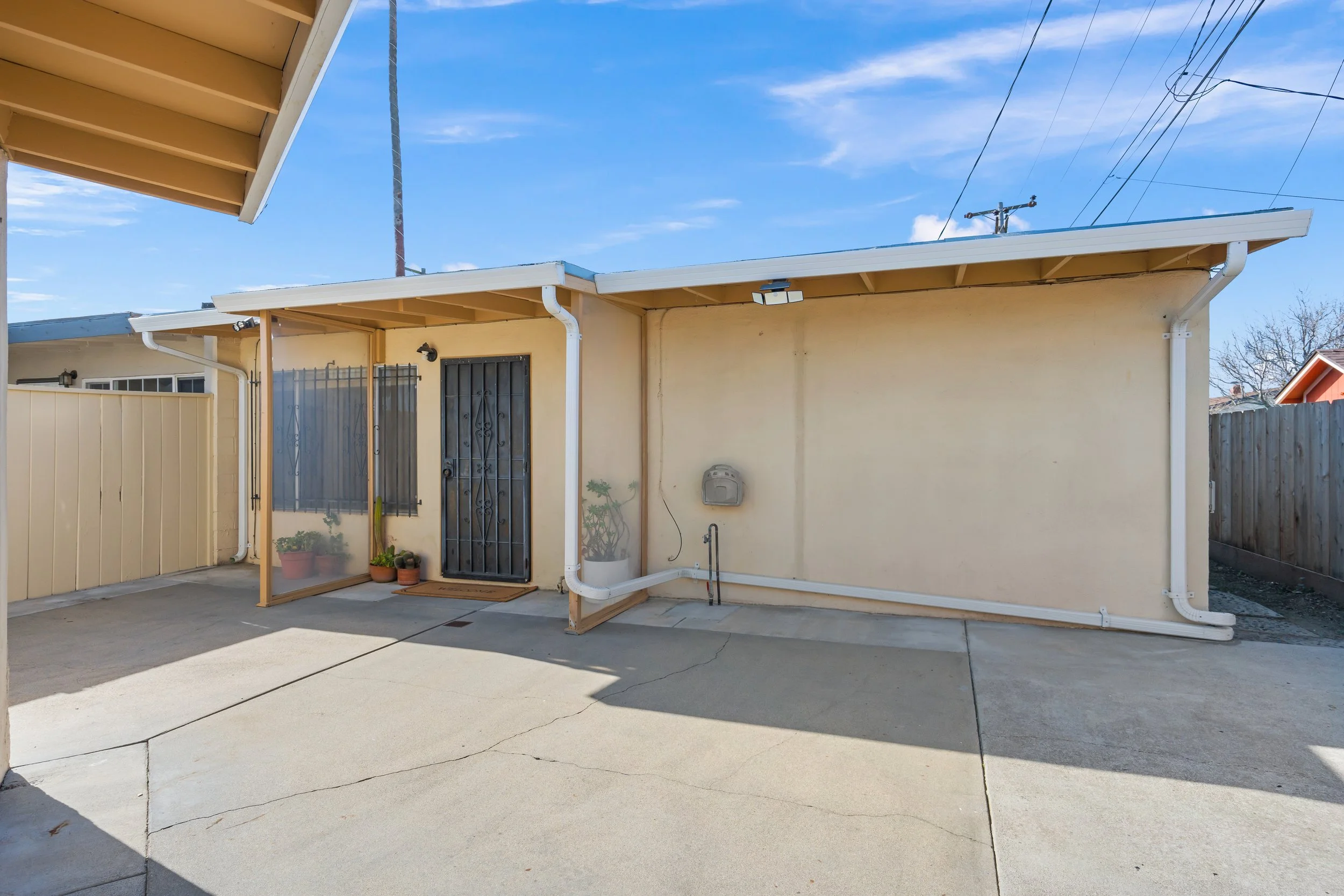 Backyard patio area with a yellow wall, concrete floor, potted plants, and a door with a security gate. Overhead roof with gutter, and a wooden fence on the sides. Clear blue sky with a few clouds in Salinas, Monterey county, CA.