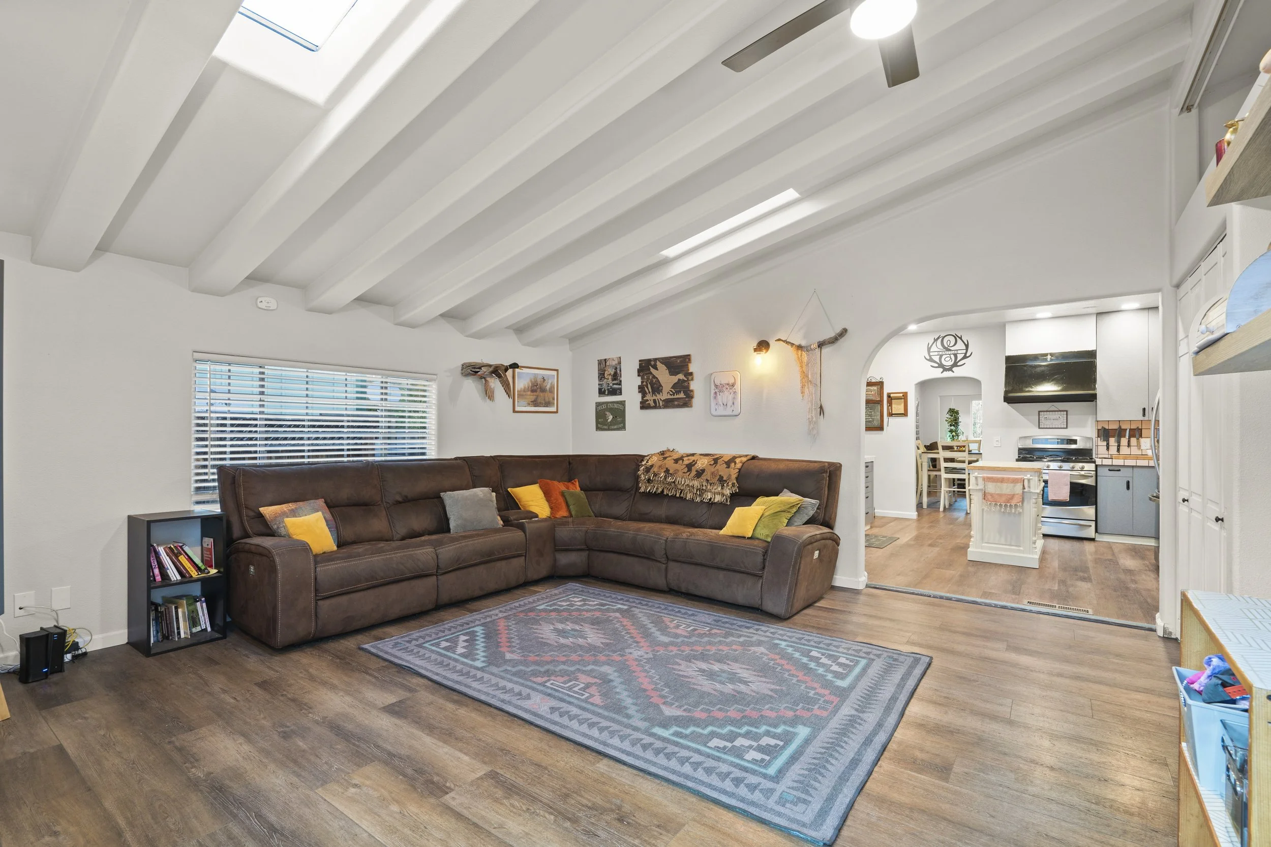 Living room with brown leather sectional, colorful pillows, patterned rug, window with blinds, wall decor, and an open view to the kitchen area.