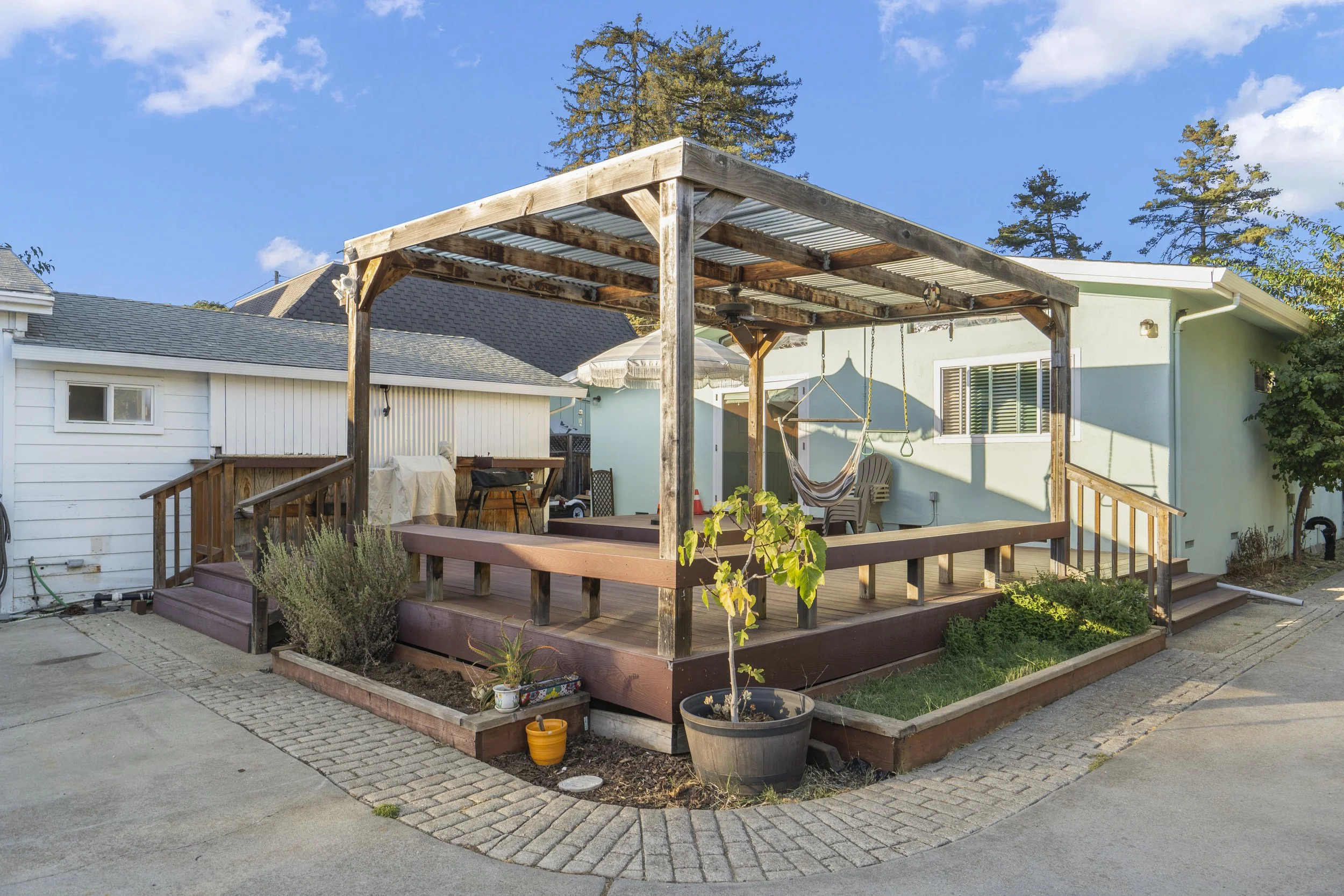 Backyard patio with wooden deck, a hammock, potted plants, and outdoor furniture under a pergola with a blue house wall in the background.