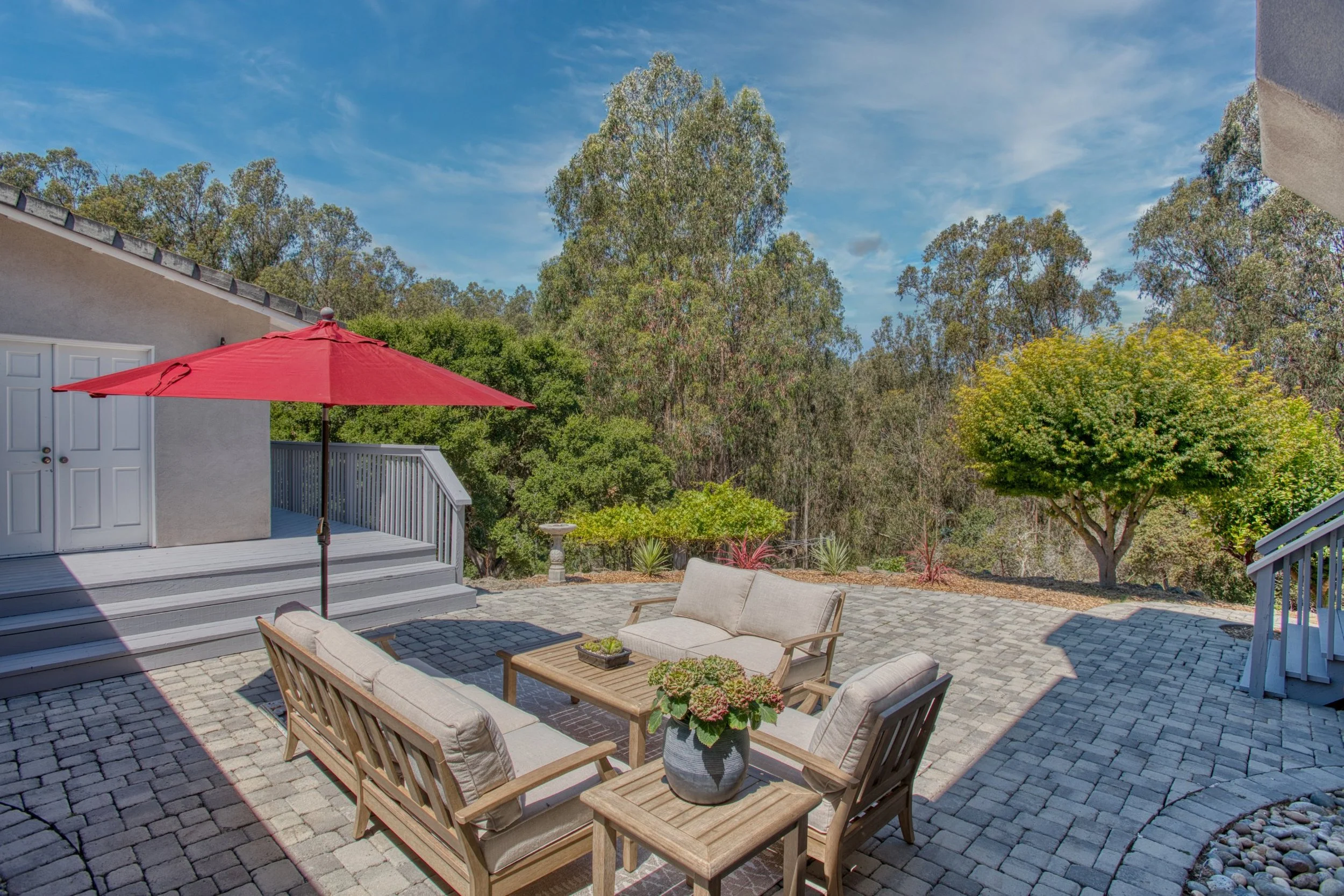 An outdoor patio area with a brick paver surface, garden furniture including a seating set with cushions and a small table, a red patio umbrella, and lush green trees and shrubs in the background under a partly cloudy sky 1650 Andreas Estates, Royal