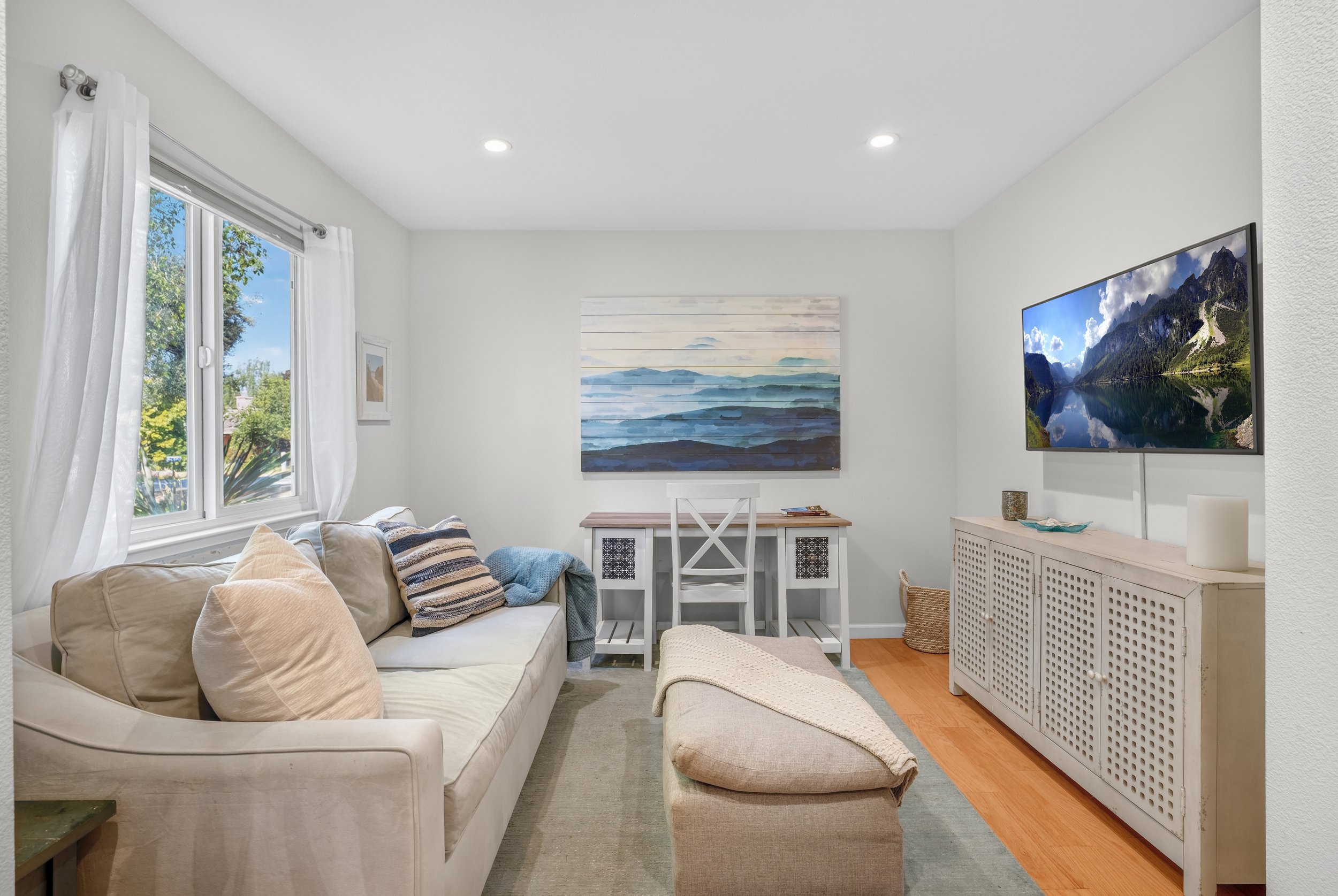 Cozy living room with white walls, a beige sofa with pillows, a window with white curtains, a small desk with a chair, a wall-mounted TV showing a mountain landscape, and wall art depicting a landscape, with hardwood and carpeted flooring.