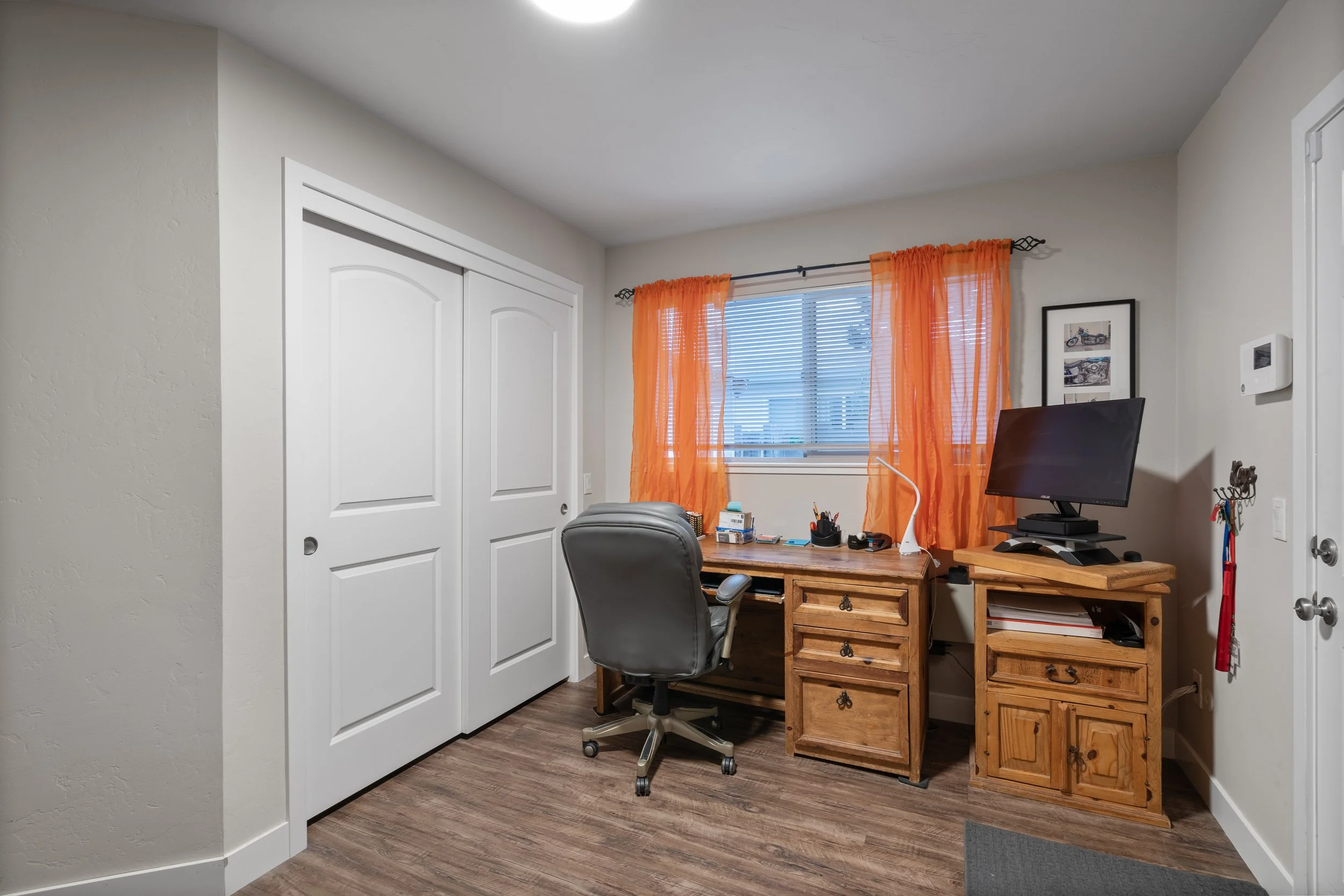 Home office with wooden desk, office chair, computer monitor, and orange curtains over window.