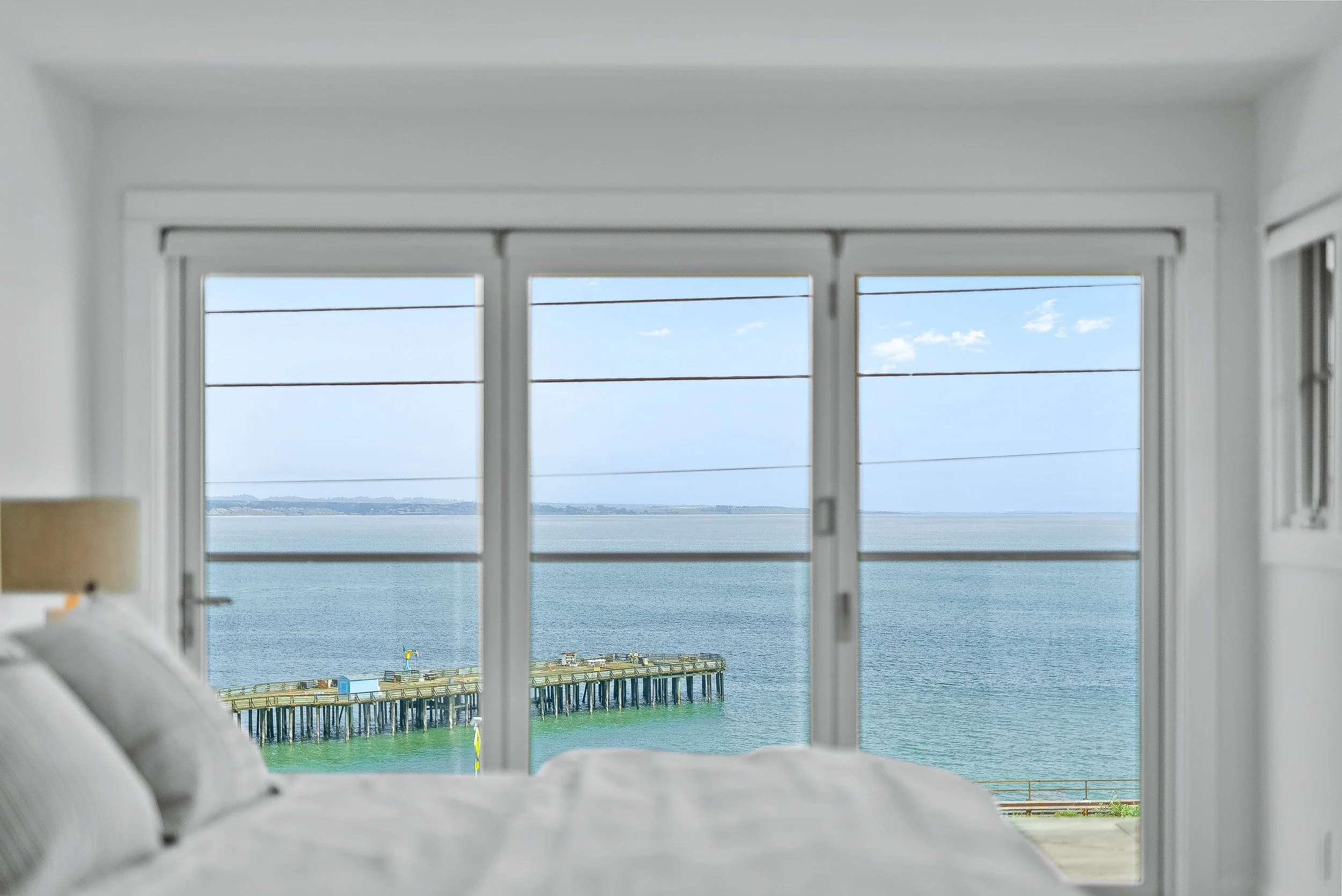 Ocean view through large sliding glass doors from a bedroom with white walls and a bed in the foreground in Capitola, California.
