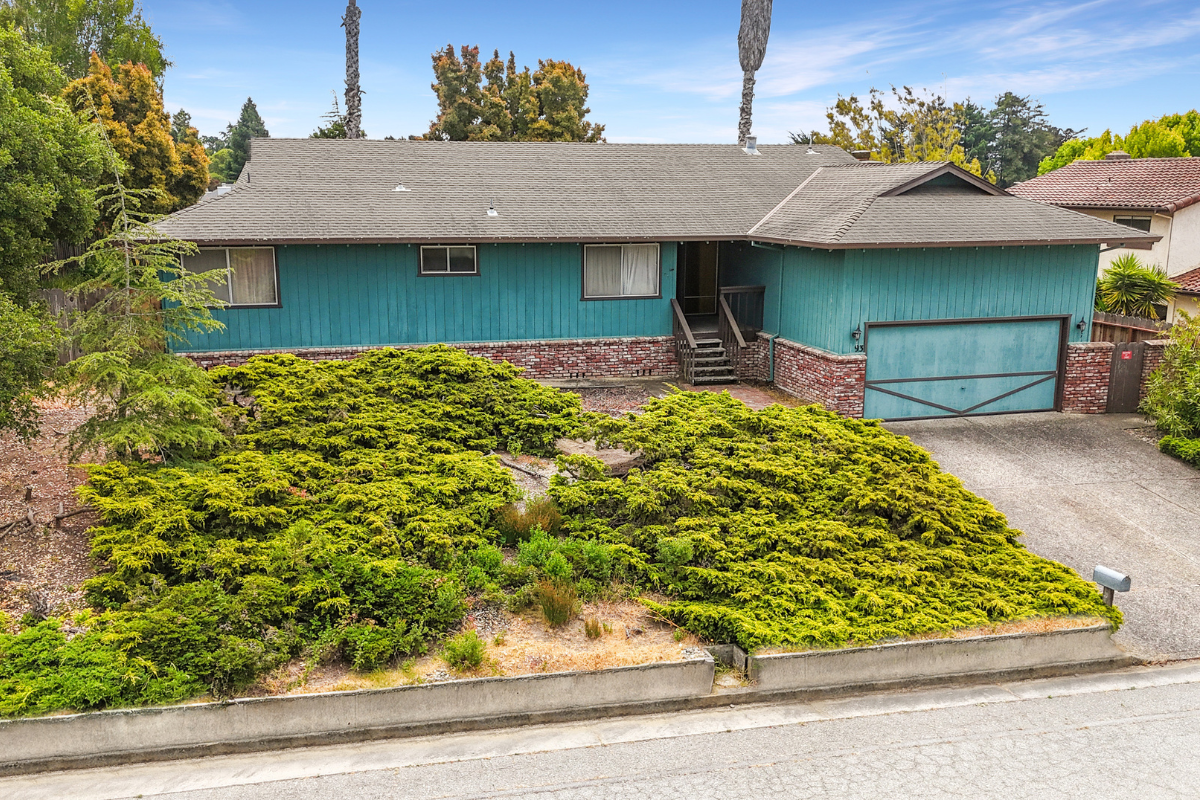 A teal-colored house with a brick foundation, a driveway, and a variety of green bushes and trees in front.