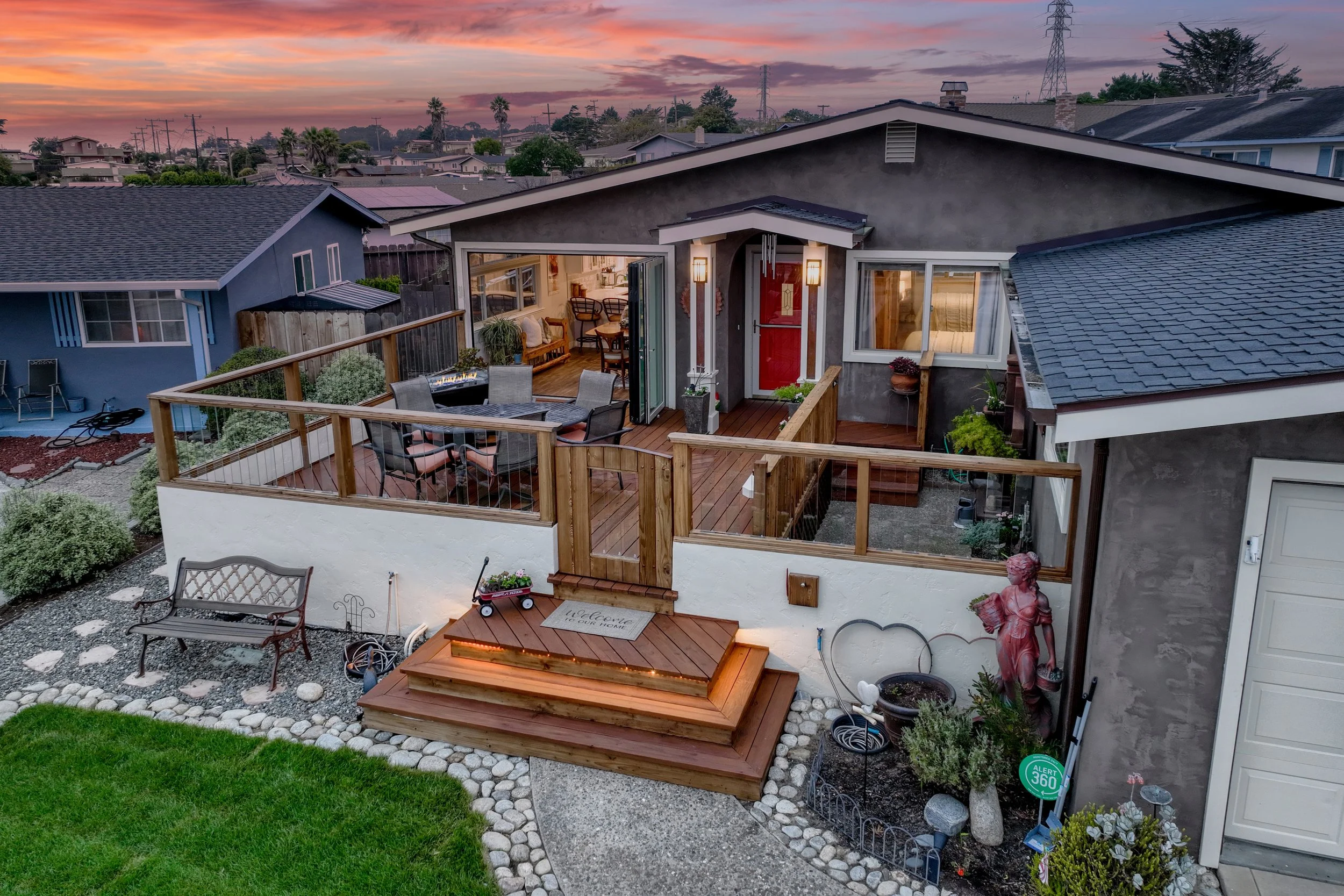 A cozy backyard patio at sunset with wooden deck, outdoor dining table, garden decor, and open sliding glass door revealing the interior of a home in Seaside, California.