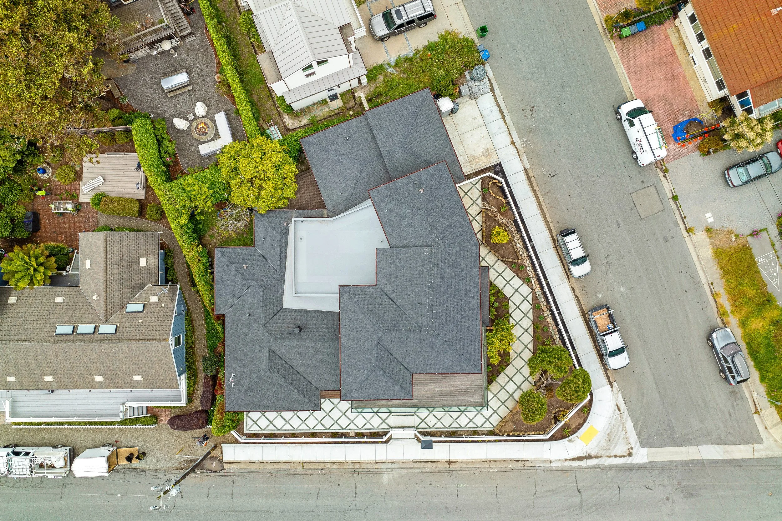Aerial view of a residential property with a gray roof, landscaped yard, adjacent driveway, and street with parked cars in Capitola.