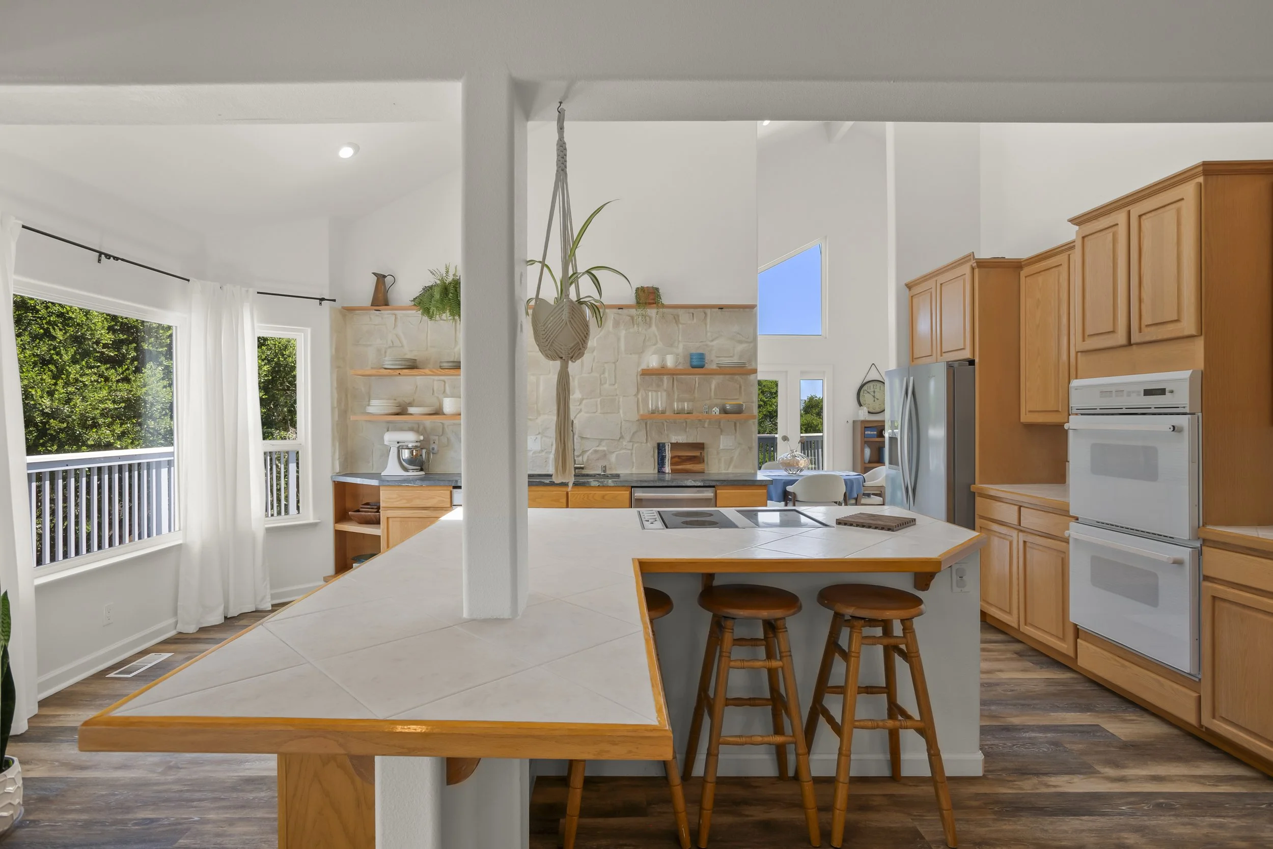 Bright kitchen with white walls, wooden cabinets, a white tiled island with wooden trim, and open shelves with dishes, a plant hanging from the ceiling, large windows with white curtains, and a wooden floor.