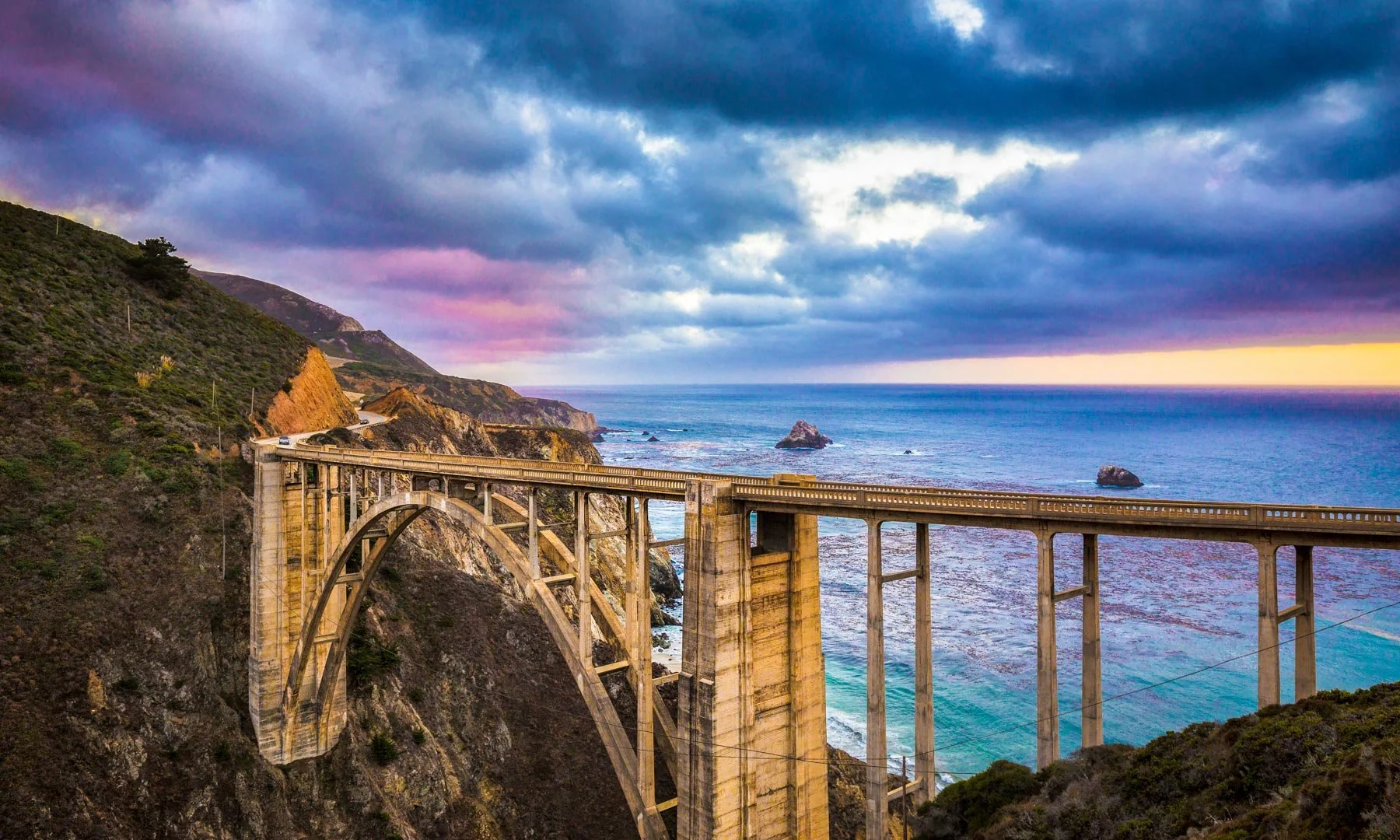 Scenic view of the Bixby Creek Bridge on the California coast at sunset, with clouds and ocean in the background.