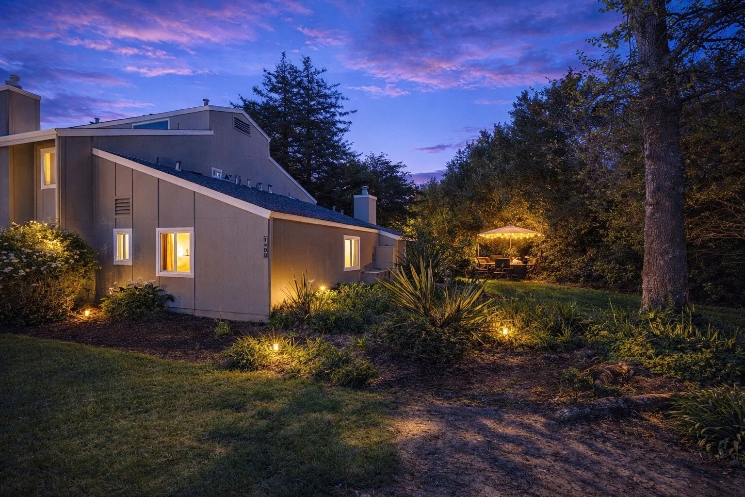 A house with illuminated windows and glowing outdoor lights at dusk, surrounded by trees and a garden with a patio and umbrella in the backyard.
