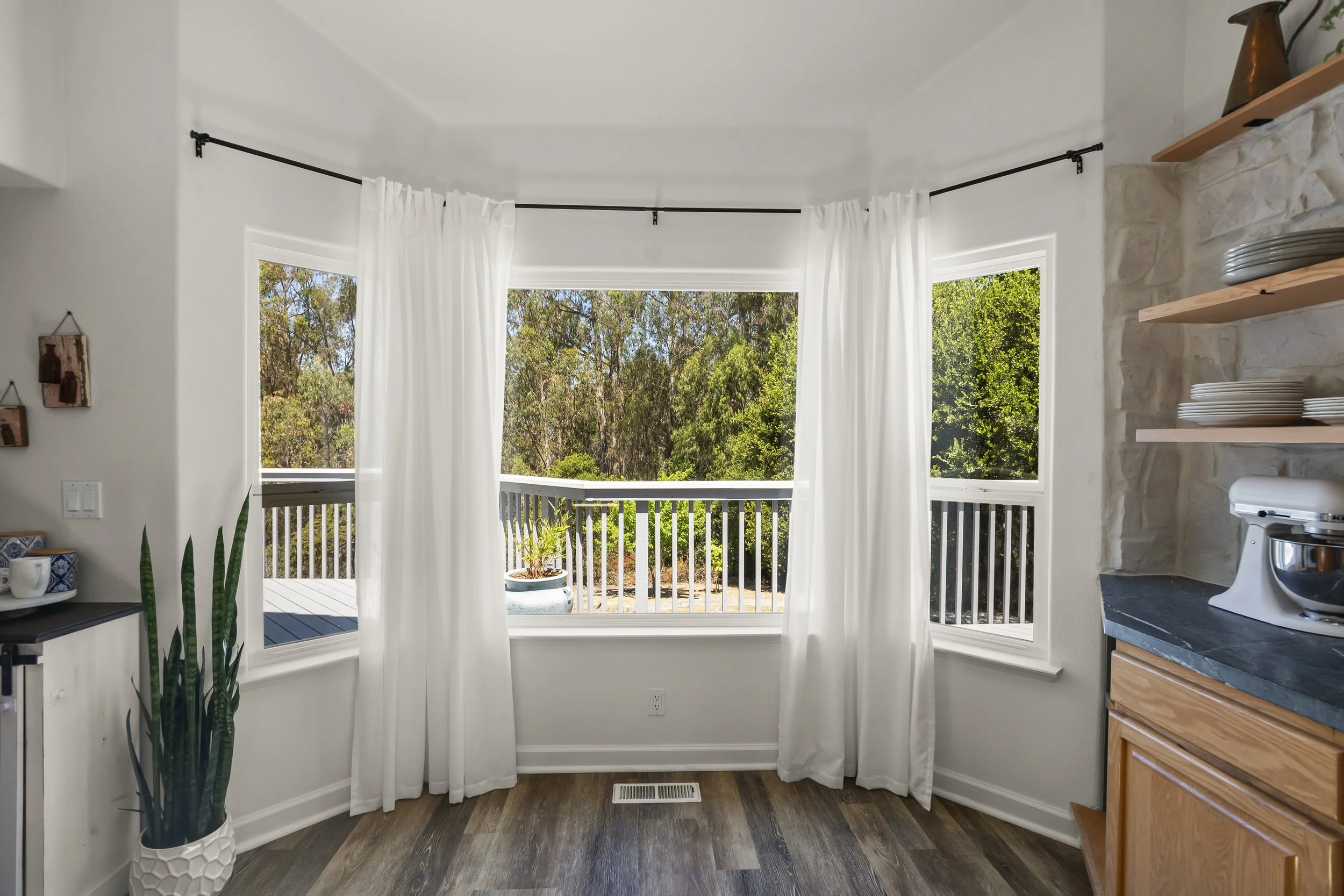 Bright kitchen corner with white curtains, wooden floor, potted plant, and a view of a deck and trees outside.