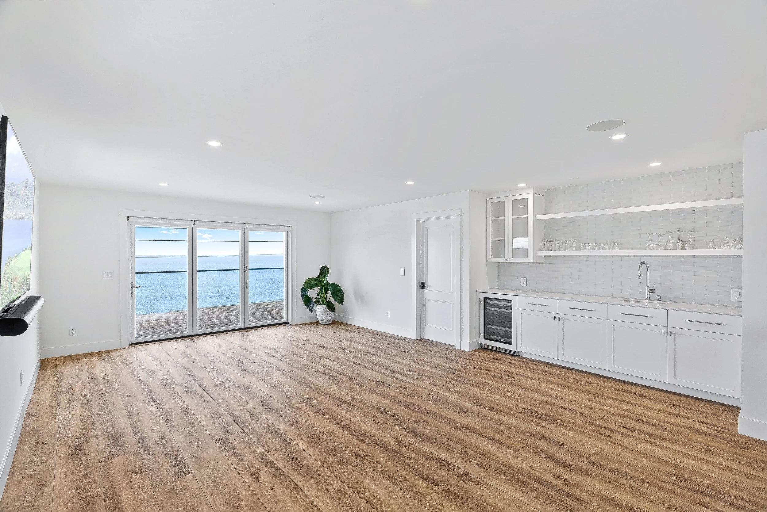 Empty living room with large sliding glass doors overlooking a body of water, wooden floors, and a white kitchen area with shelves and a wine cooler in Capitola, California.