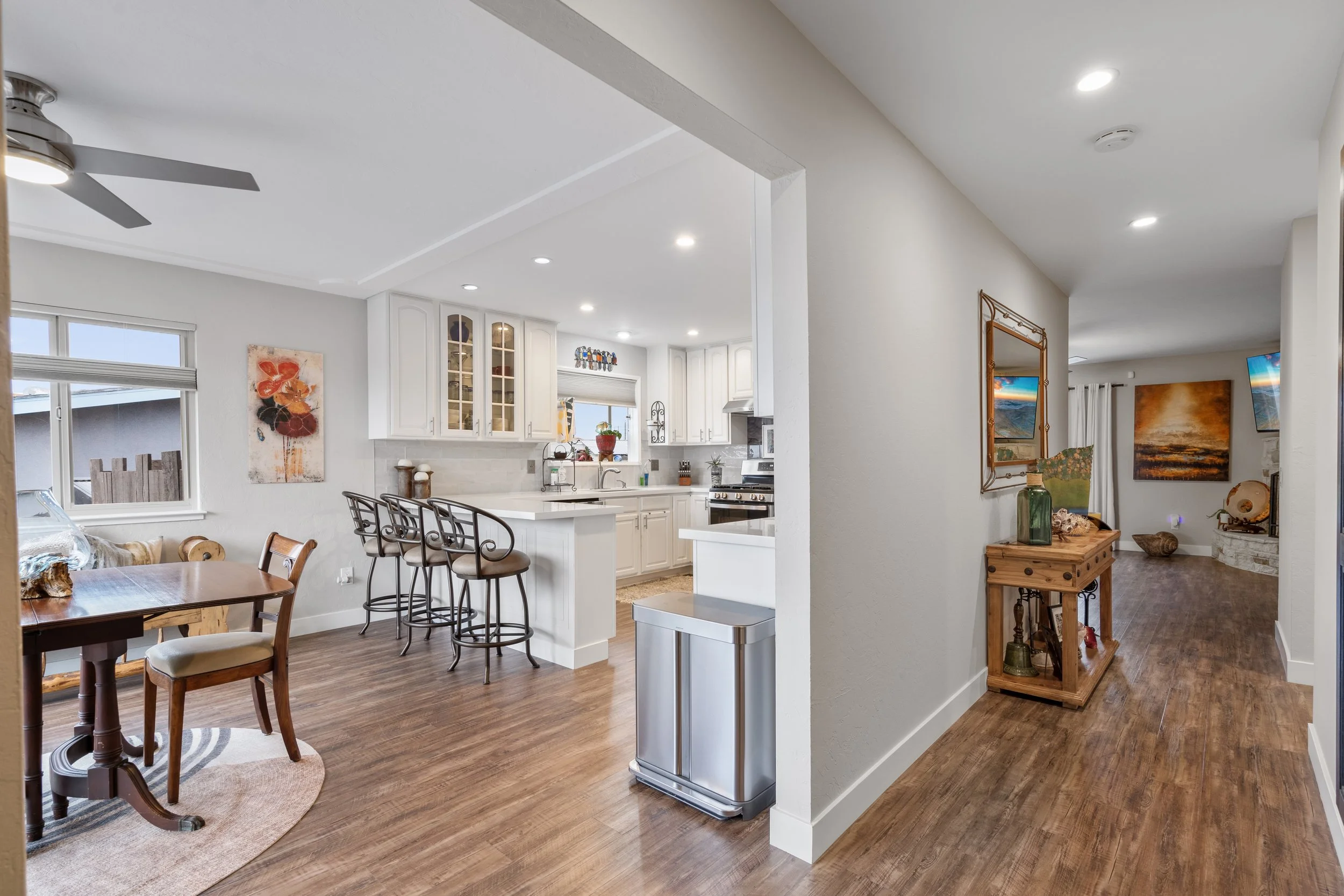 Open-concept kitchen and dining area with wooden floors, white cabinets, and bar stools, connected to a living room with artwork and television.