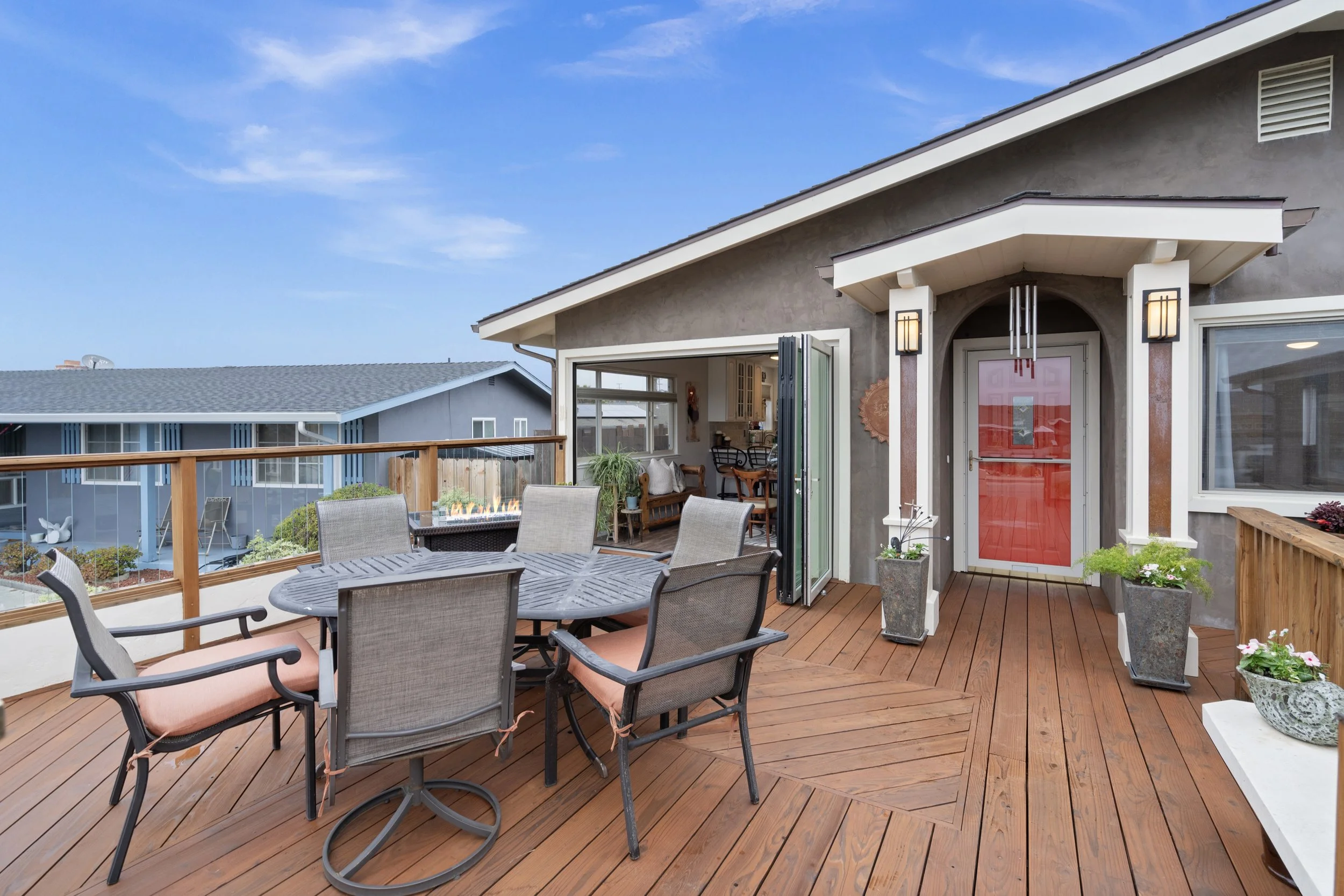 Outdoor wooden deck with a round table and six chairs, potted plants near a red front door, and an open view into a house interior with plants and furniture, under a blue sky.