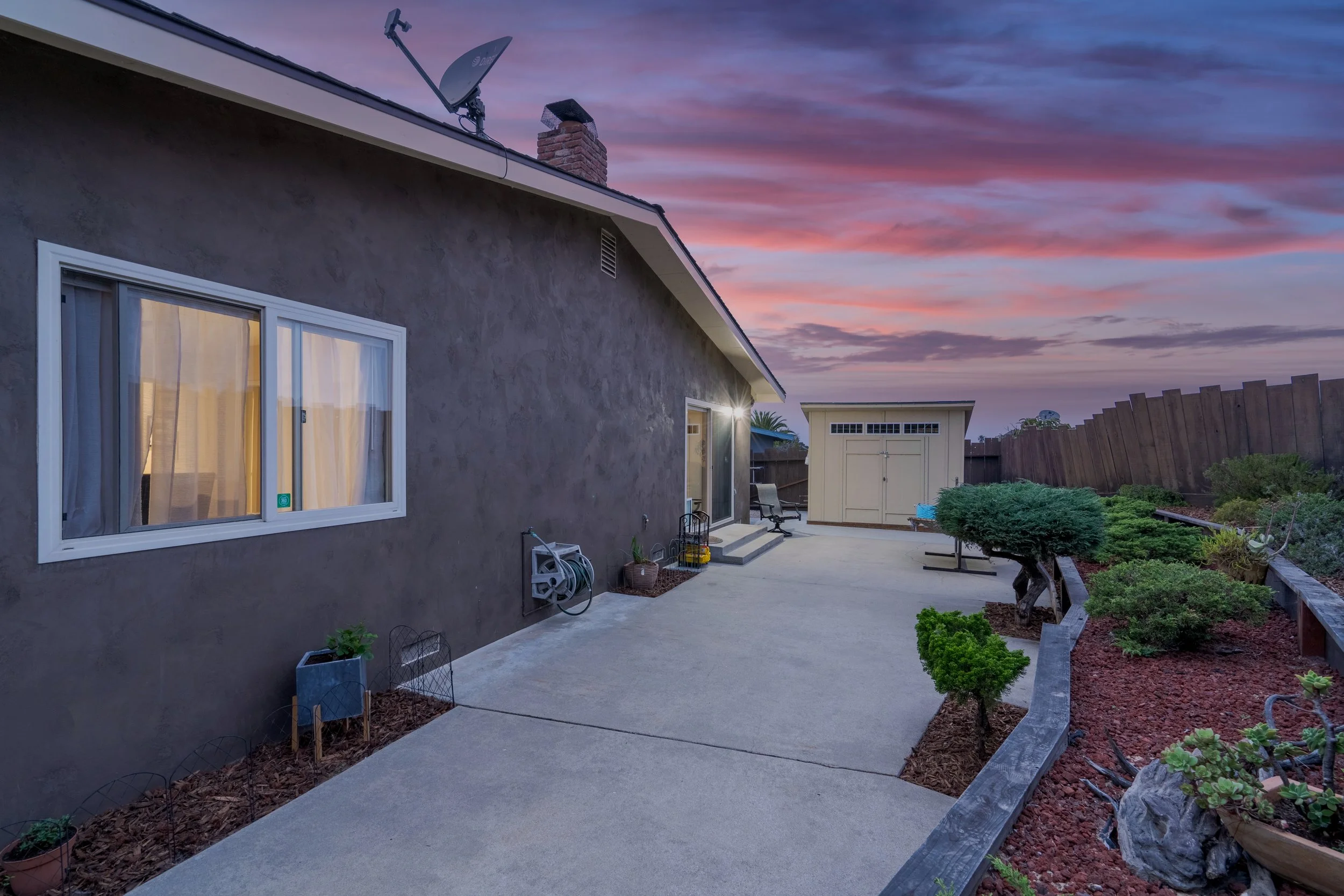 Backyard patio at sunset with a concrete area, a garden bed with plants, a shed, and a dark-colored house wall with a window.