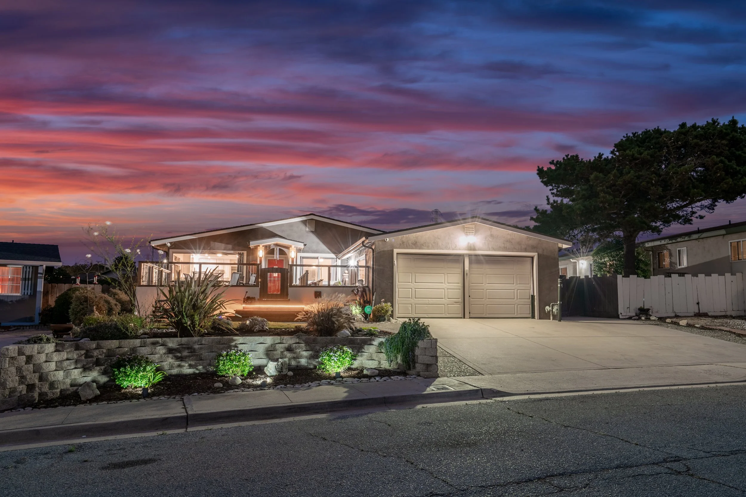 A suburban house at dusk with a colorful sunset sky, lit exterior lights, landscaped front yard, and a two-car garage in Seaside, California.