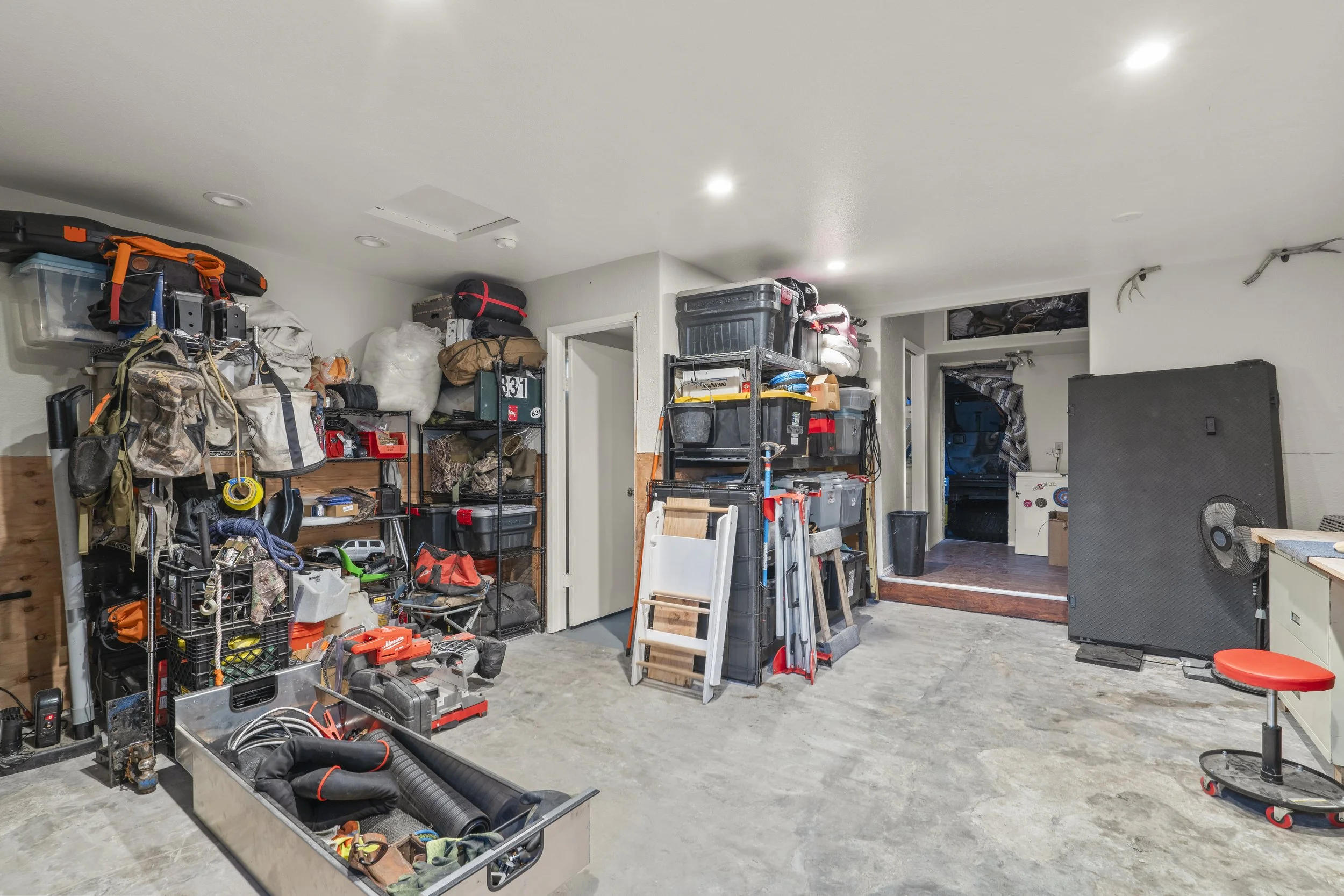A cluttered garage storage area with shelves and bins filled with camping gear, tools, and supplies, with an open doorway leading to another room.