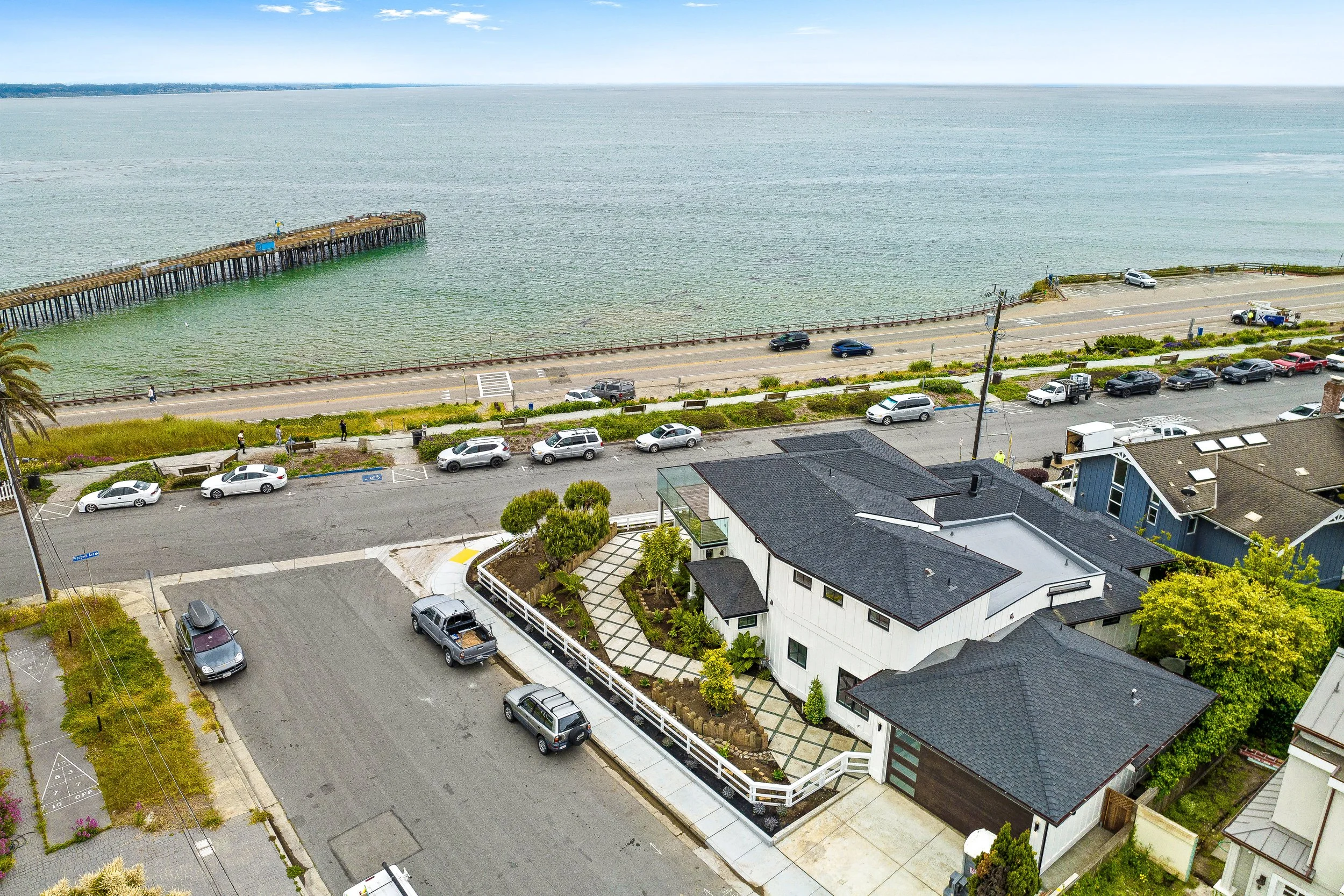 Aerial view of a coastal neighborhood with houses and a parking lot, overlooking a beach and ocean pier, under a partly cloudy sky.