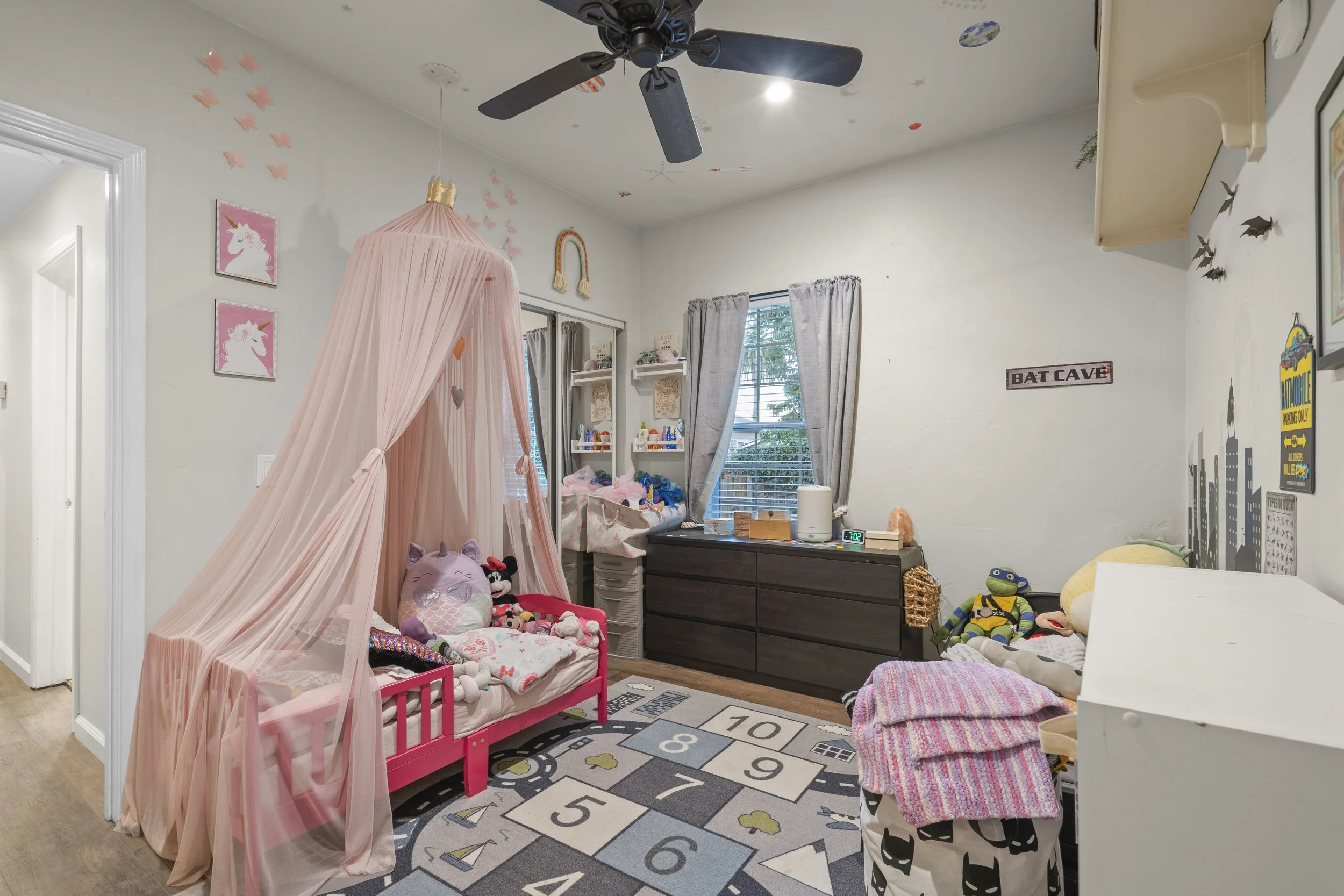 Children's bedroom with pink princess-themed bed, canopy, stuffed toys, and colorful decor, including wall art and toys on furniture, with a window and a black dresser.
