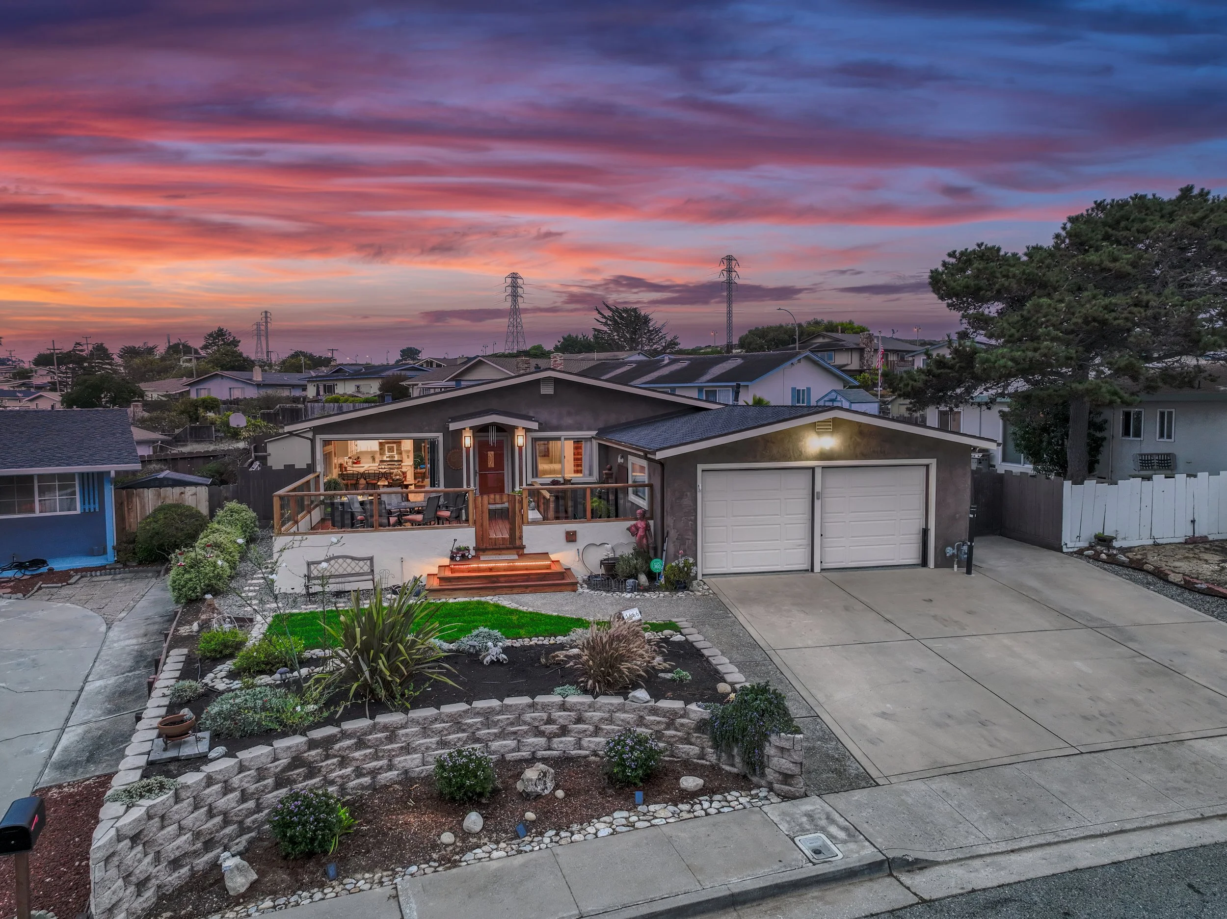 A front view of a single-story house at sunset with a well-lit interior, a small front porch with seating, a manicured yard with various plants and a brick retaining wall, and a large driveway leading to a double garage. The sky is colorful with shad