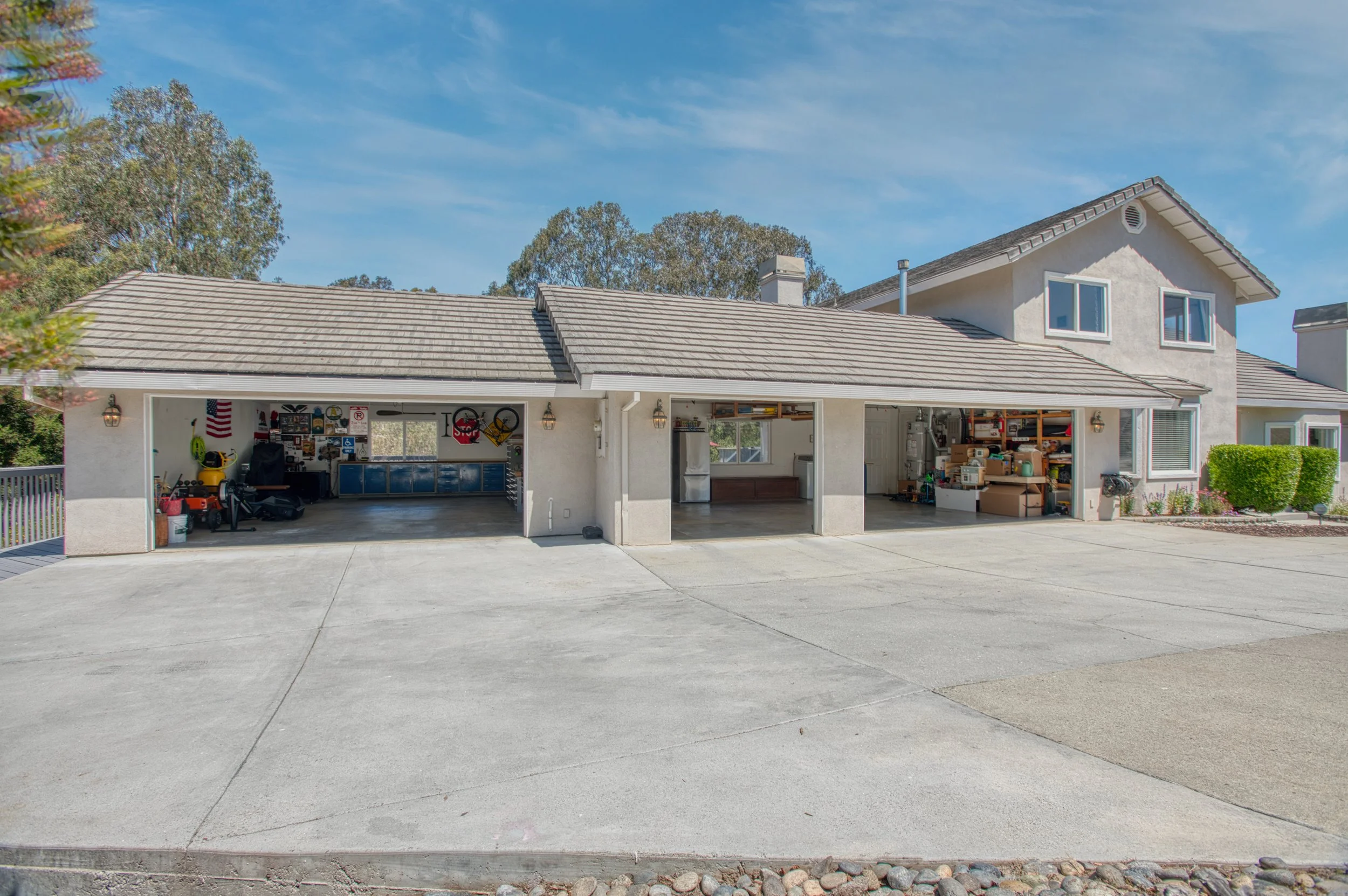 A three-car garage with open doors, filled with tools and storage, attached to a two-story house with a gray roof, white walls, and a small front yard with shrubs, under a blue sky.