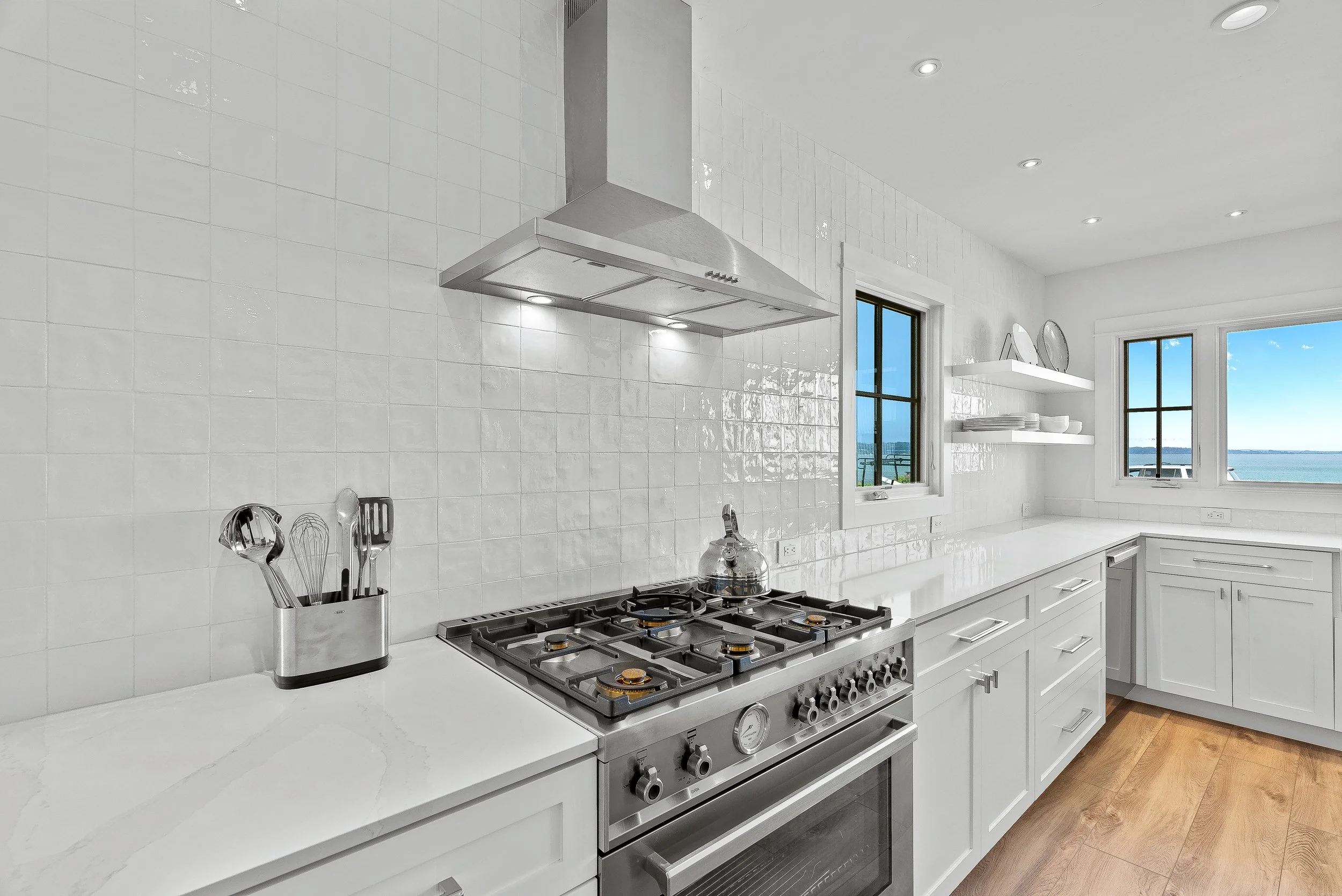 Modern white kitchen with stainless steel stove, white cabinets, open shelving with dishes, and windows showing a waterfront view in Capitola, California.